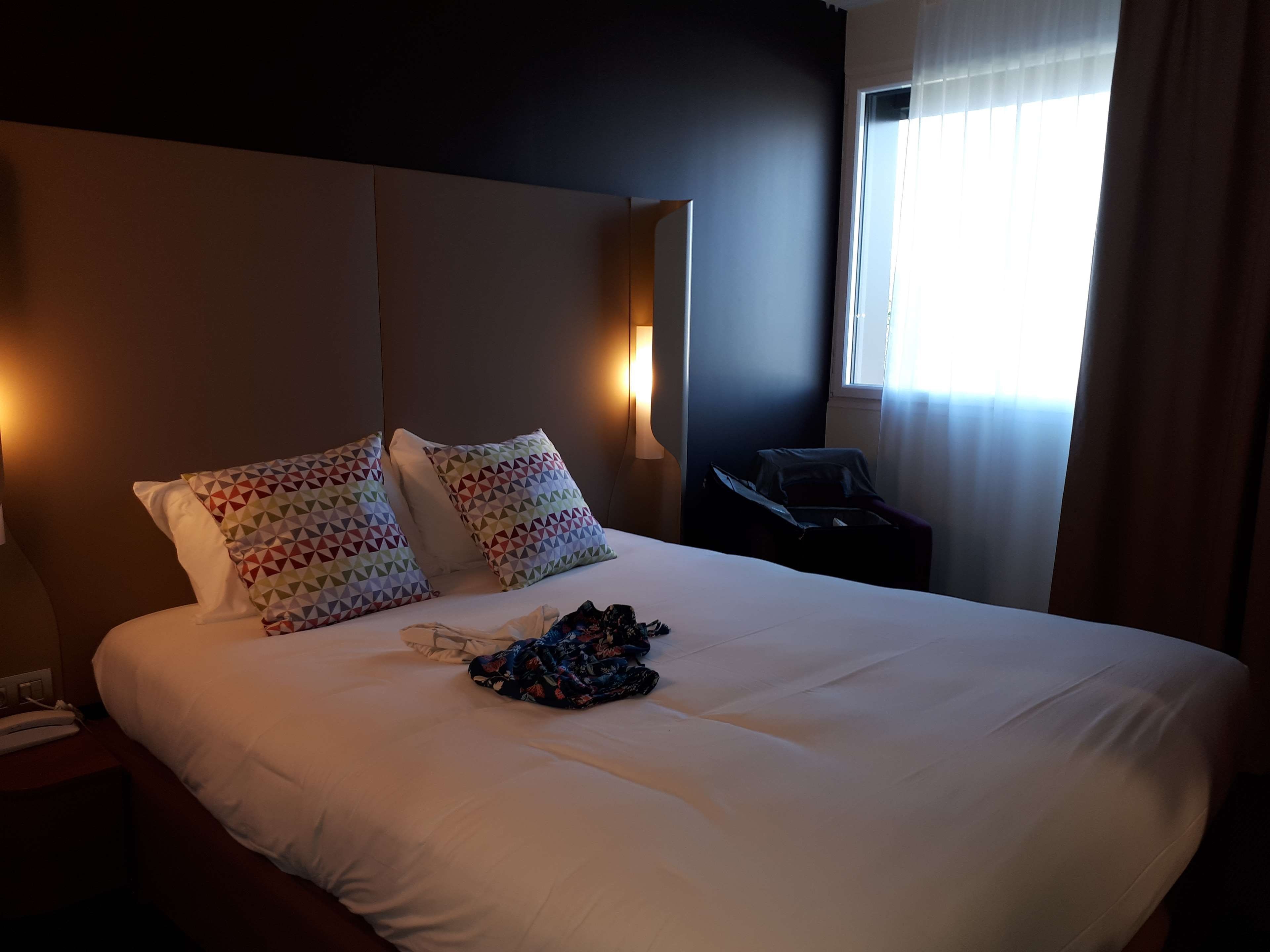 A hotel room with a large bed, two patterned pillows, a bedside lamp, a suitcase, and a window with curtains.