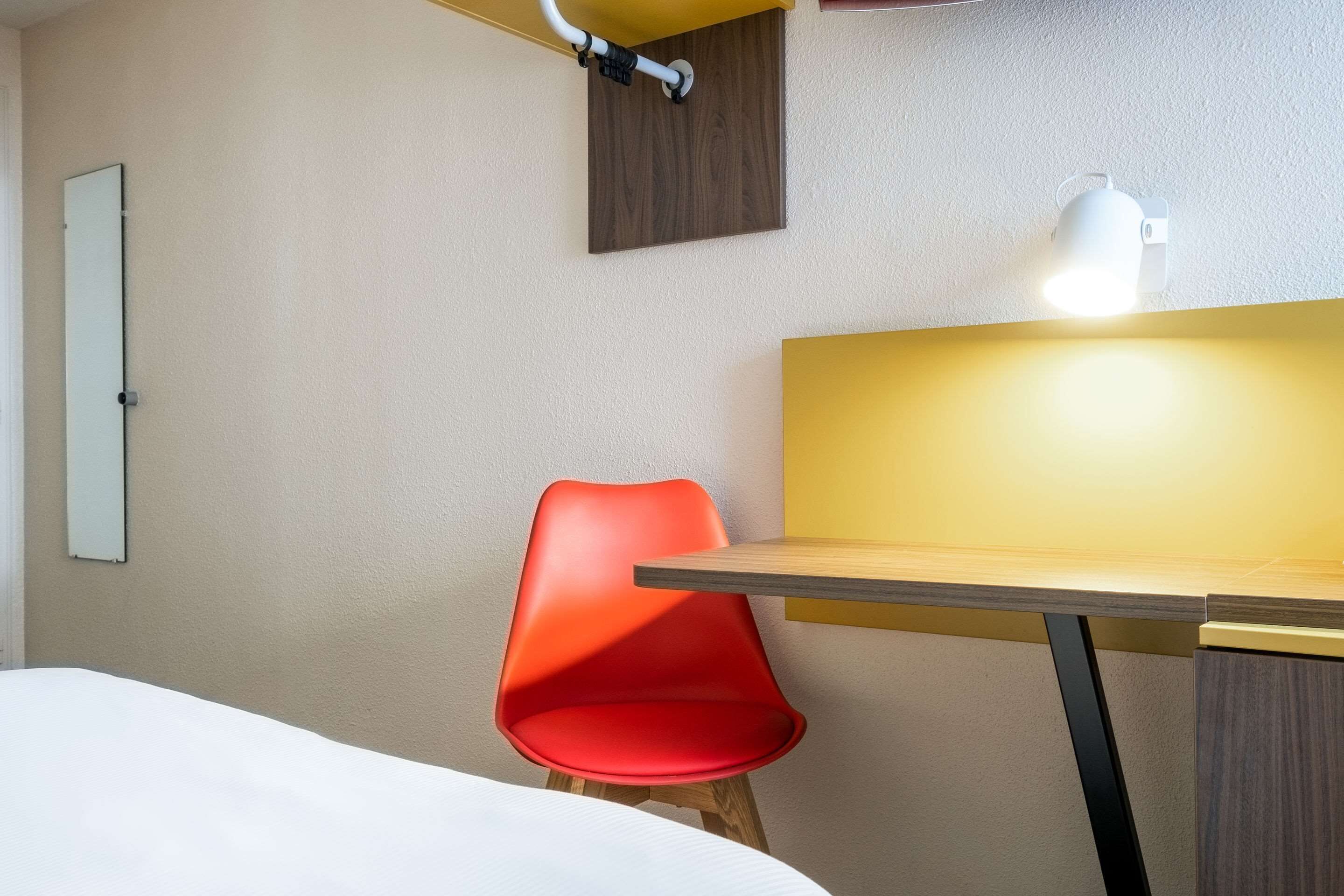 Ett hotellrum med en säng, en röd stol, ett skrivbord i trä, en vit lampa och en liten hylla i trä.