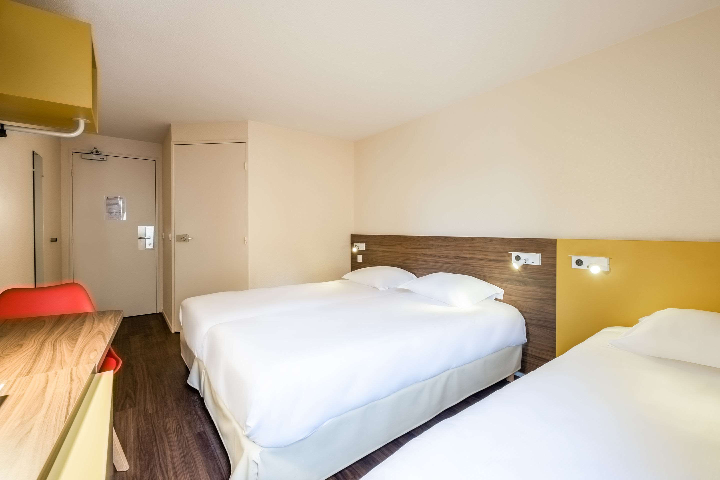 A hotel room with two beds, a wooden desk, a red chair, and a door.