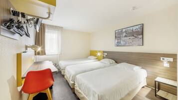A hotel room with a desk, two beds, a red chair, a window with curtains, a wall-mounted lamp, and a framed picture.