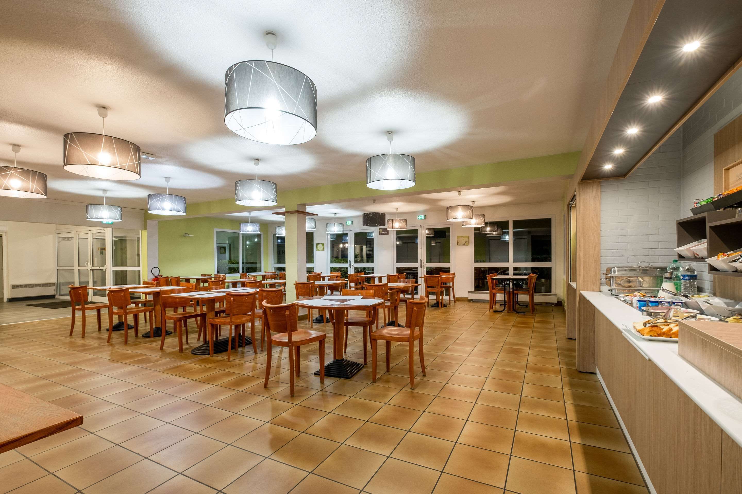 Un grand espace dédié à la salle à manger, avec plusieurs tables et chaises, des luminaires au plafond et un comptoir de cuisine où sont disposés des aliments.