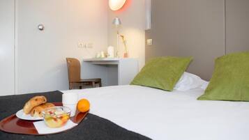 A hotel room with a bed, green pillows, a desk with a lamp, a chair, and breakfast items on a tray.