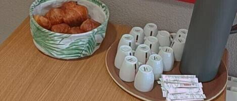 A small wooden table with a bowl of croissants, a plate with white mugs, and a black tumbler.