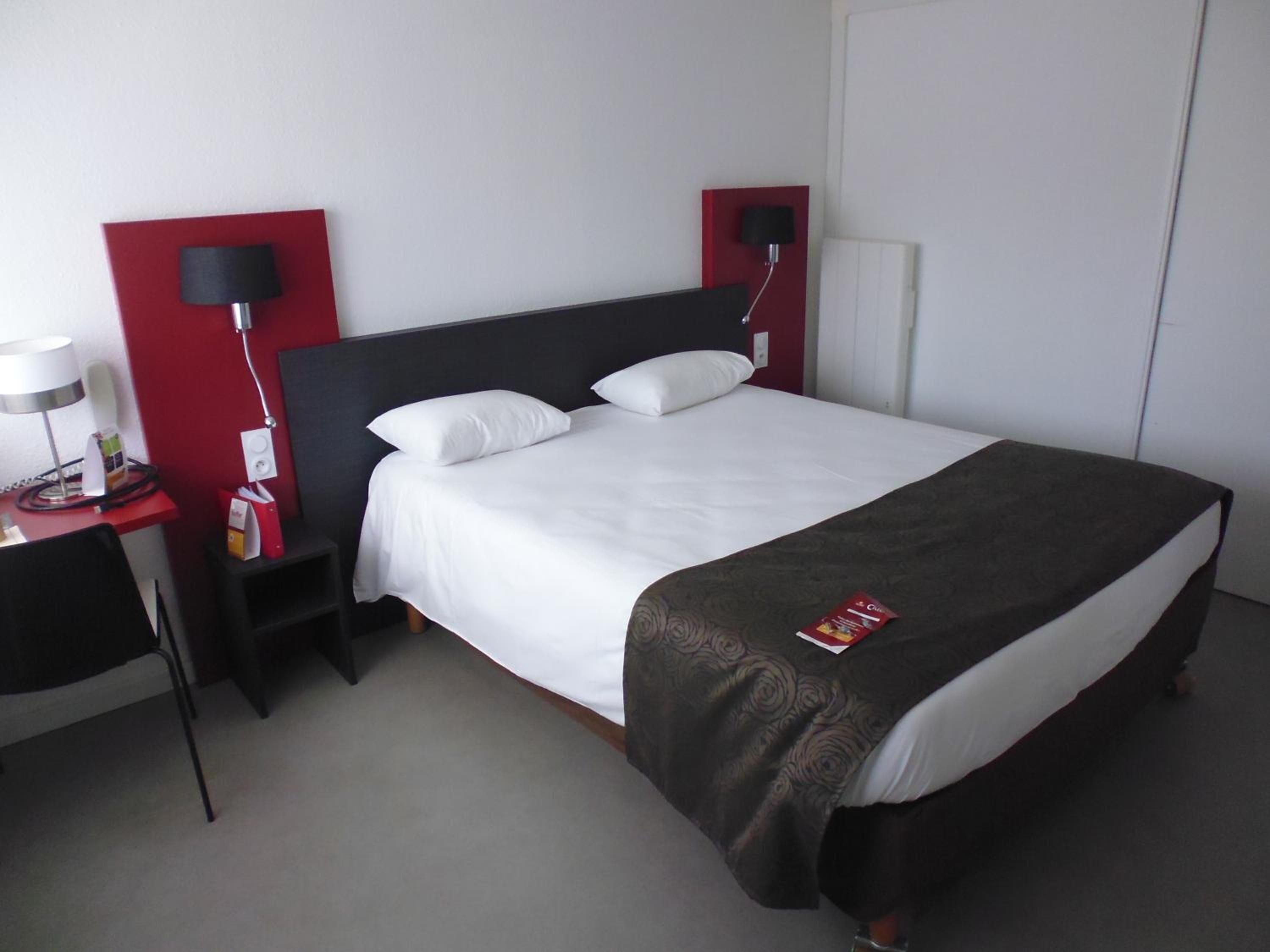 A hotel room with a bed, a desk, a chair, a lamp, and a red accent wall.