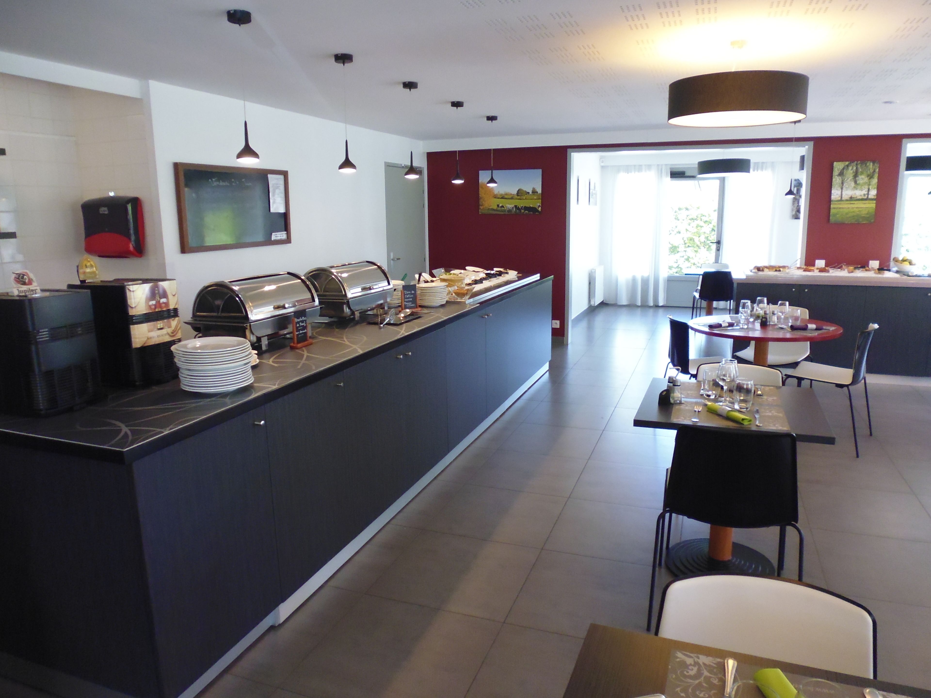 Une cuisine d’hôtel moderne, dotée de buffets, d’un espace repas avec tables et chaises, et offrant une vue sur l’extérieur grâce à de grandes fenêtres.