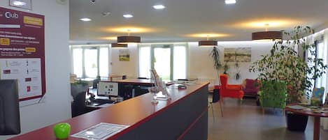 Zona de recepción de hotel con un mostrador, computadoras y asientos. Hay plantas y un cuadro en las paredes.