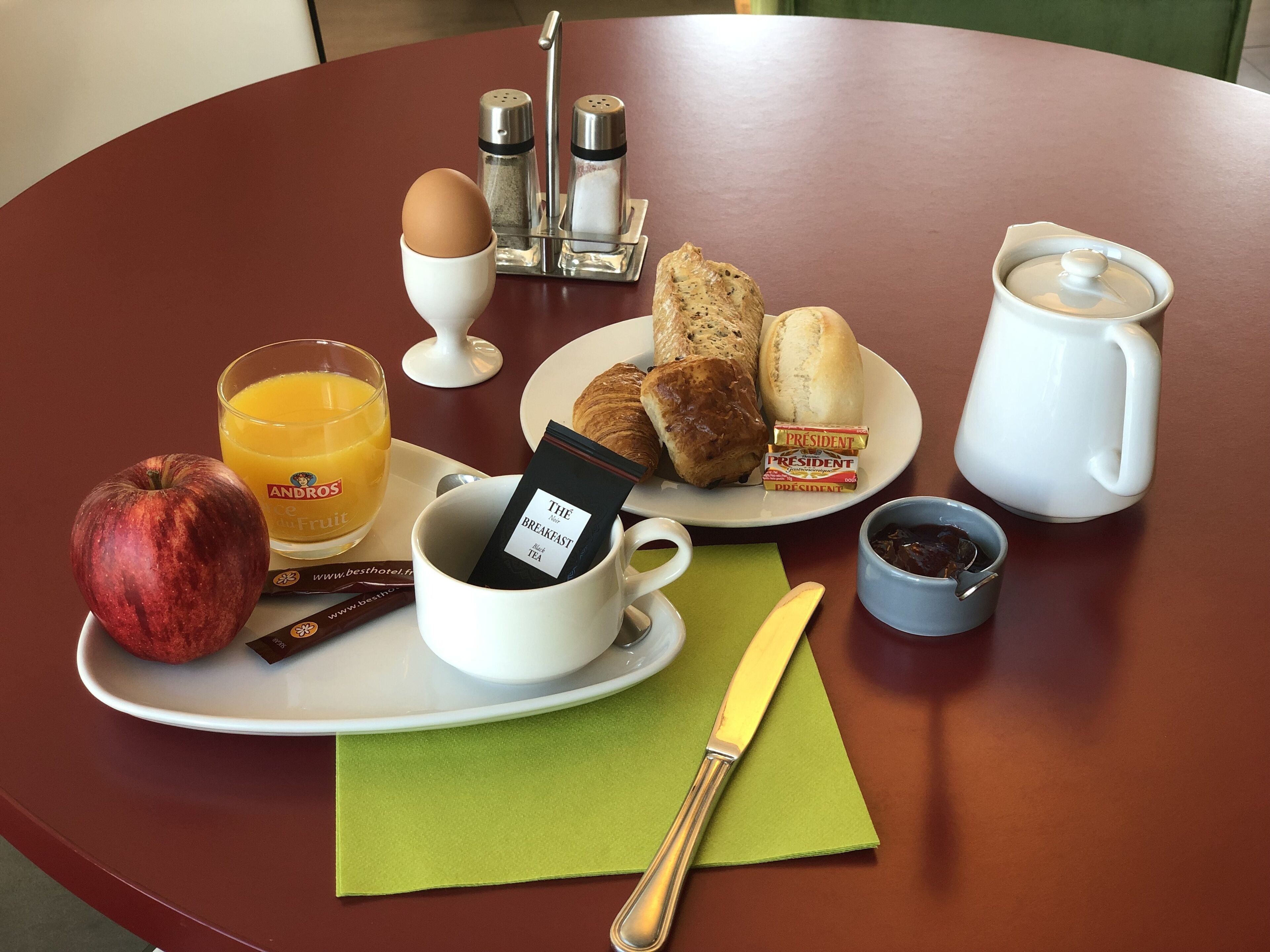 A breakfast tray with a croissant, a muffin, a coffee mug, a small bowl of jam, a packet of tea, a glass of orange juice, and a whole apple.
