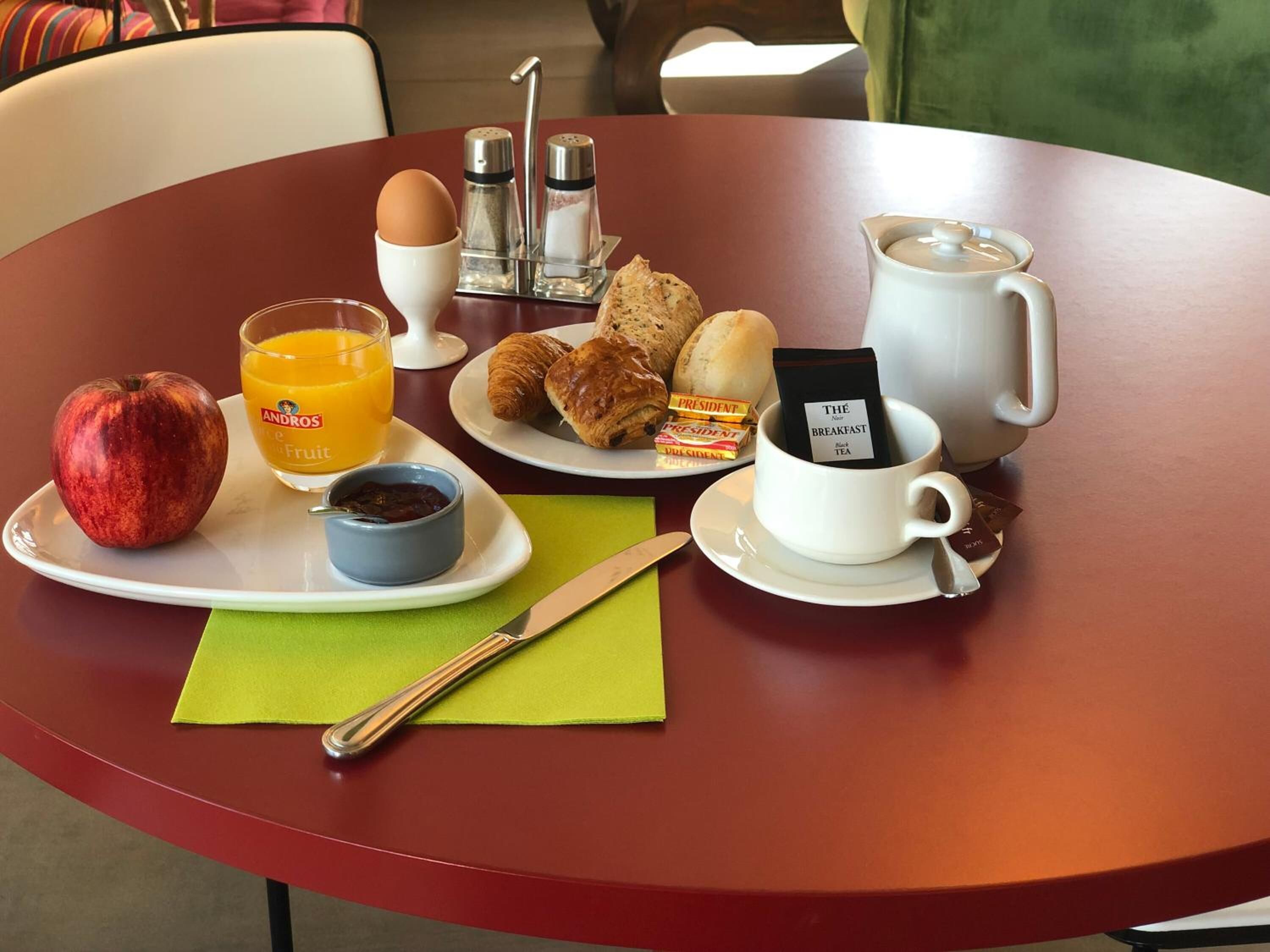 A breakfast tray with an apple, orange juice, croissants, a cup of tea, and a coffee cup on a table.