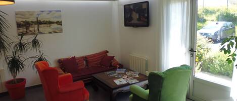 A living room with a red sofa, a green armchair, and a coffee table with magazines. There is a painting on the wall and a window with a view of a car outside.
