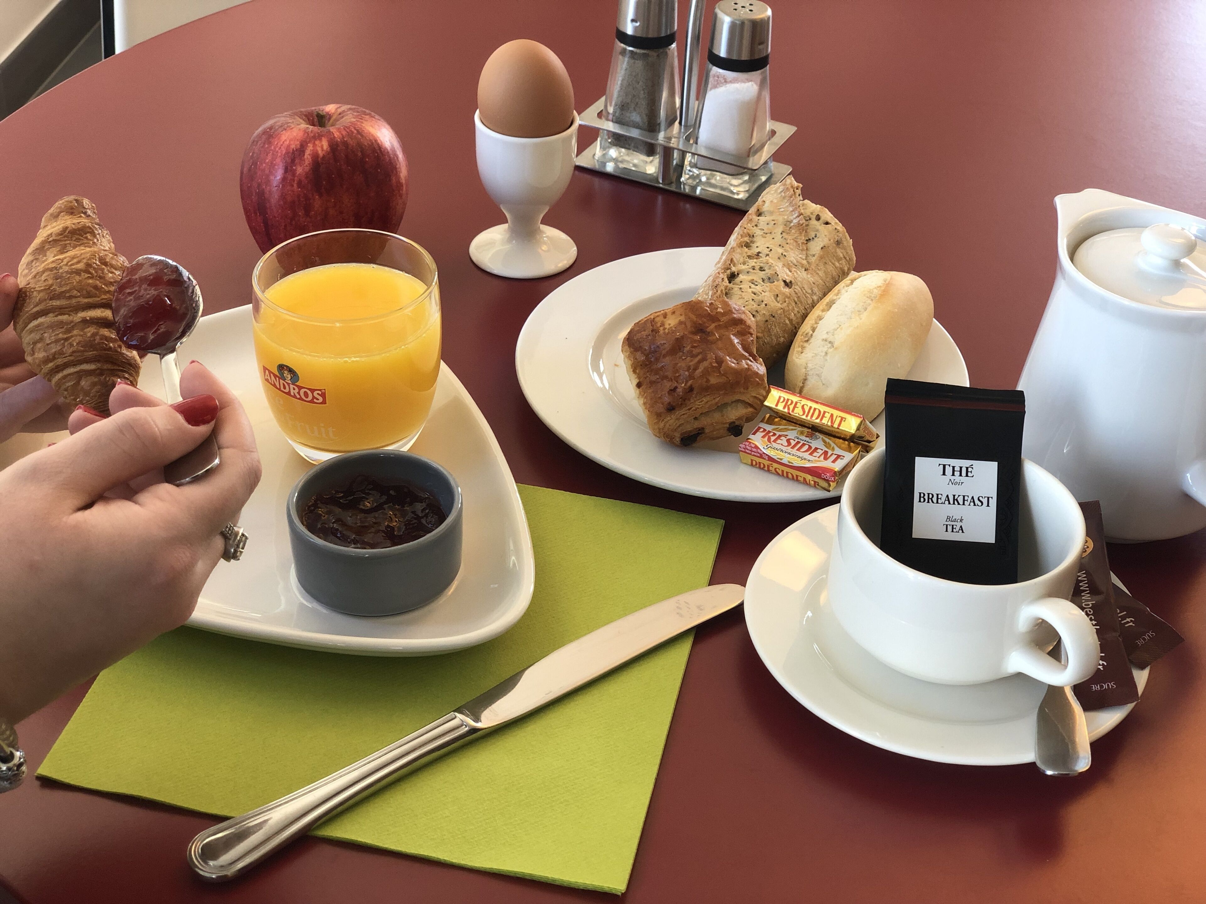 Un desayuno con croissants, una manzana, jugo de naranja, una taza de té negro, y un plato con pasteles y un sándwich.