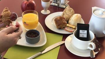 Un desayuno con croissants, una manzana, jugo de naranja, una taza de té negro, y un plato con pasteles y un sándwich.
