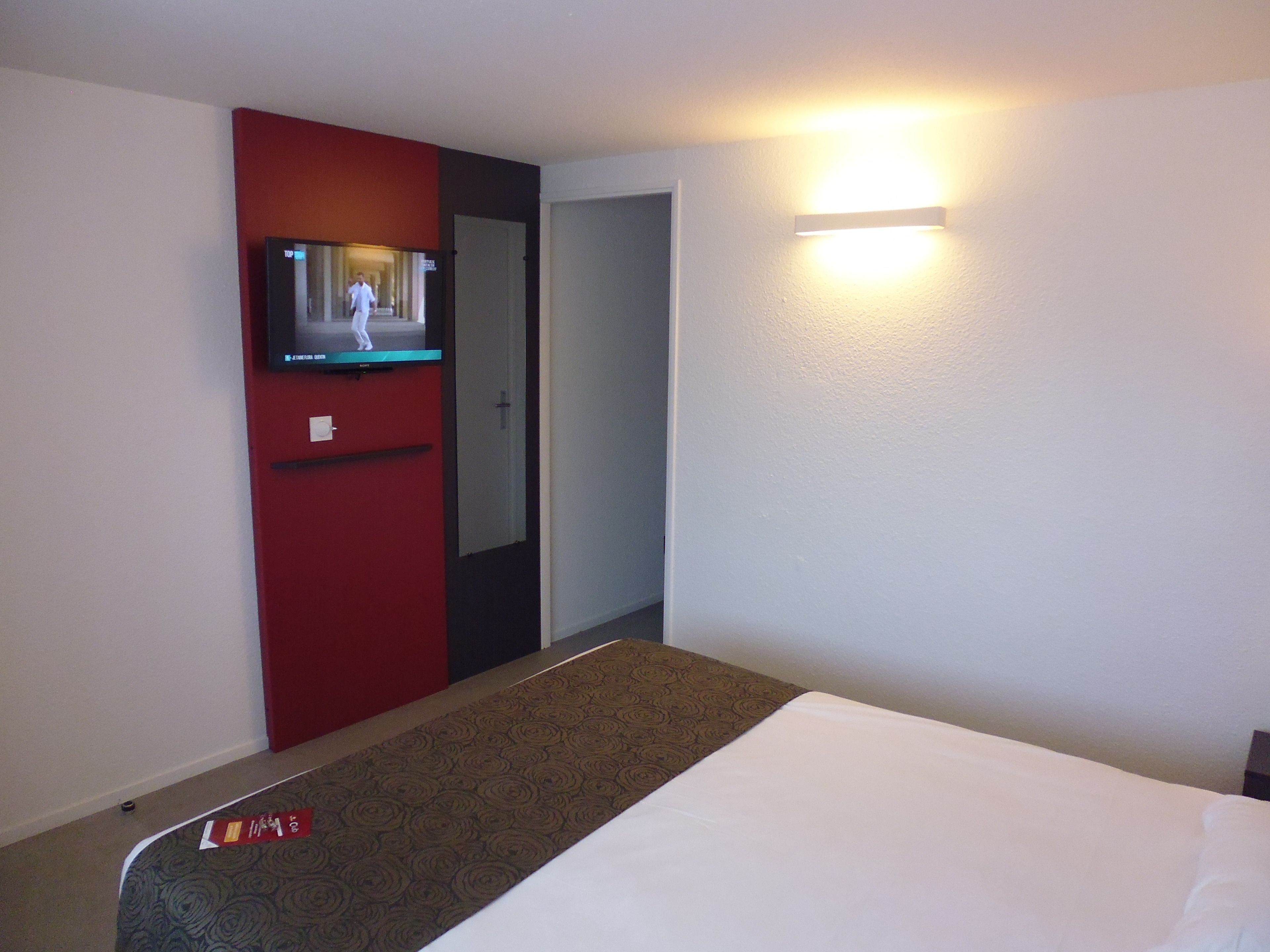 A hotel room with a bed, a television on a red cabinet, a door, and a wall-mounted light.
