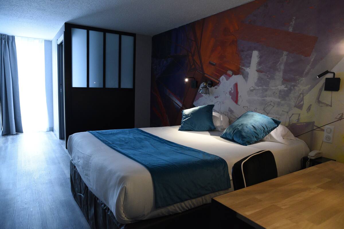 Une chambre d’hôtel comprenant un lit, un bureau, une chaise, une lampe et un mur décoré d’œuvres d’art abstraites.