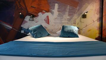 Una cama con colcha azul y dos almohadas a juego, una pared con arte abstracto, y dos lámparas de pared.