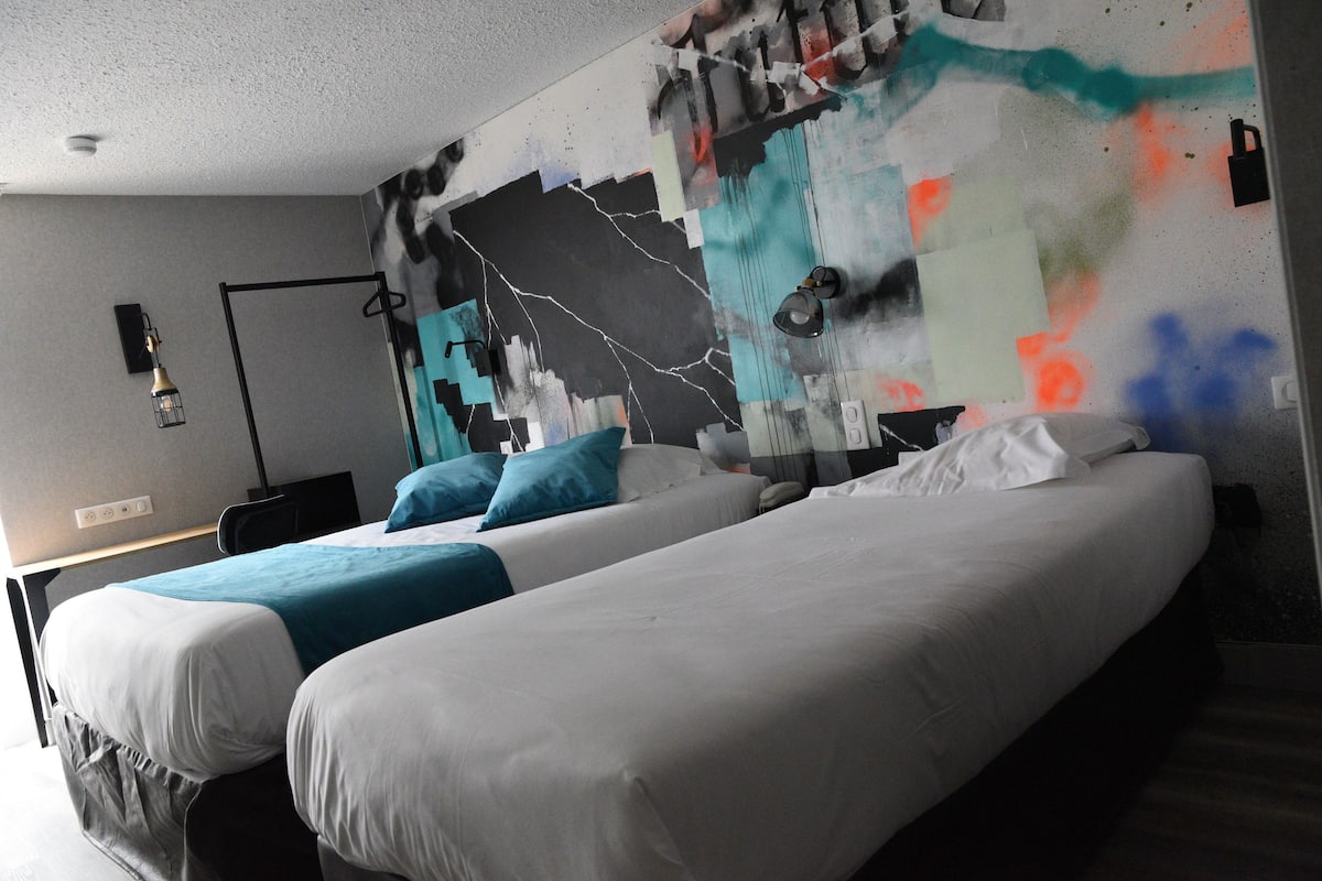 Une chambre d’hôtel avec deux lits, une fresque représentant un paysage urbain sur le mur et un bureau avec une lampe.