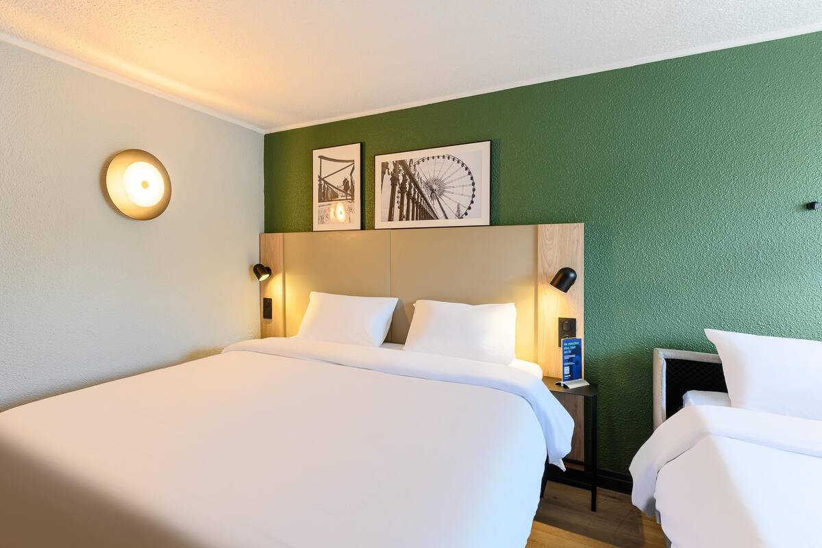 Quarto de hotel com uma cama grande, duas mesas de cabeceira com abajures, um relógio de parede e duas imagens emolduradas.