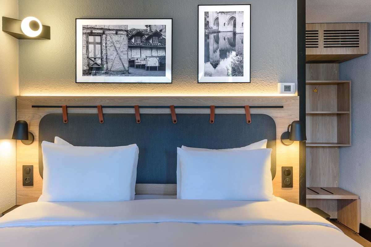 Uma cama bem arrumada com lençóis brancos, uma cabeceira com design texturizado e dois quadros na parede acima.