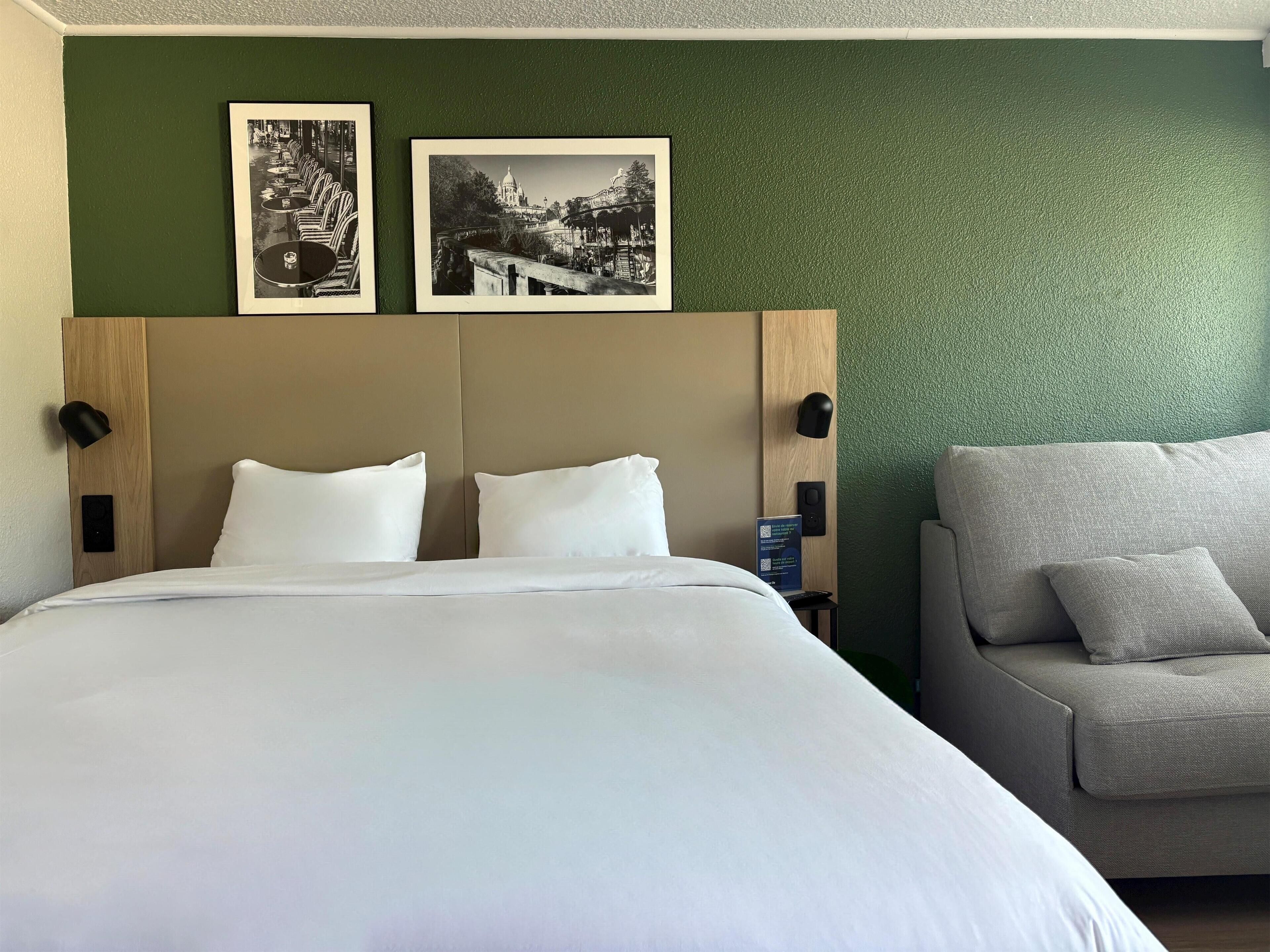 A hotel room with a large bed, a grey sofa, two framed black and white photographs on the wall, and a green textured wall.