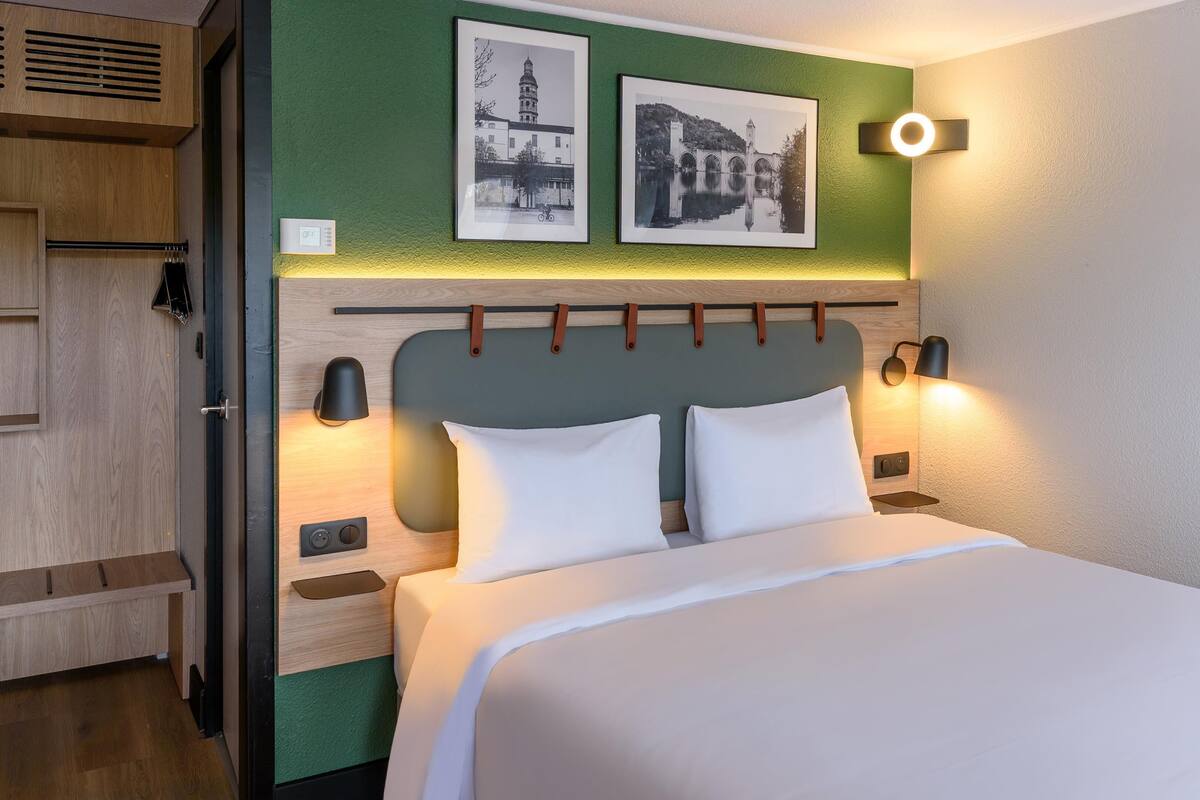 Quarto de hotel com uma cama, duas luminárias fixadas na parede, uma parede verde decorativa e duas imagens emolduradas.