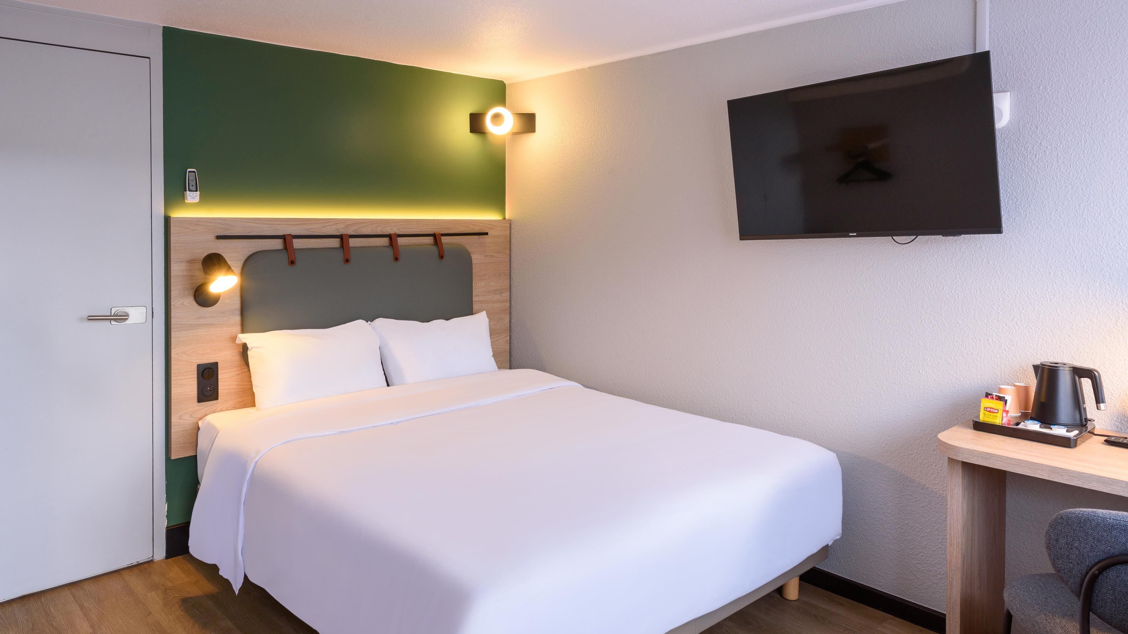 Hotellihuone, jossa on sänky, työpöytä, kahvinkeitin, televisio ja yksi seinä on maalattu vihreällä, mikä tuo huoneeseen väriä.