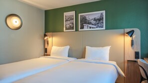 Quarto de hotel com duas camas, uma luminária redonda na parede e duas imagens emolduradas.