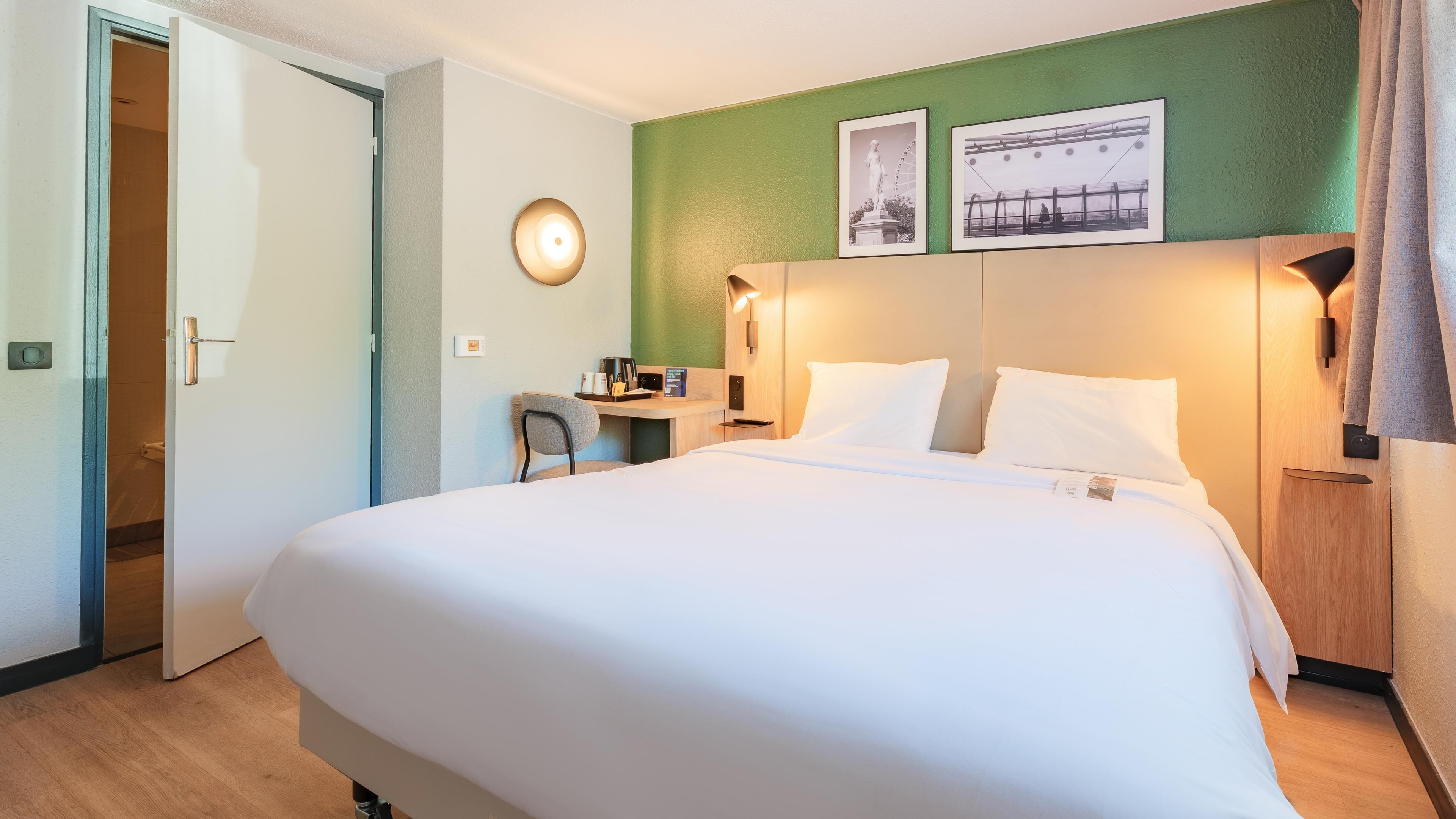 Habitación de hotel con una cama grande, un escritorio con silla, una lámpara de pared y dos cuadros enmarcados.