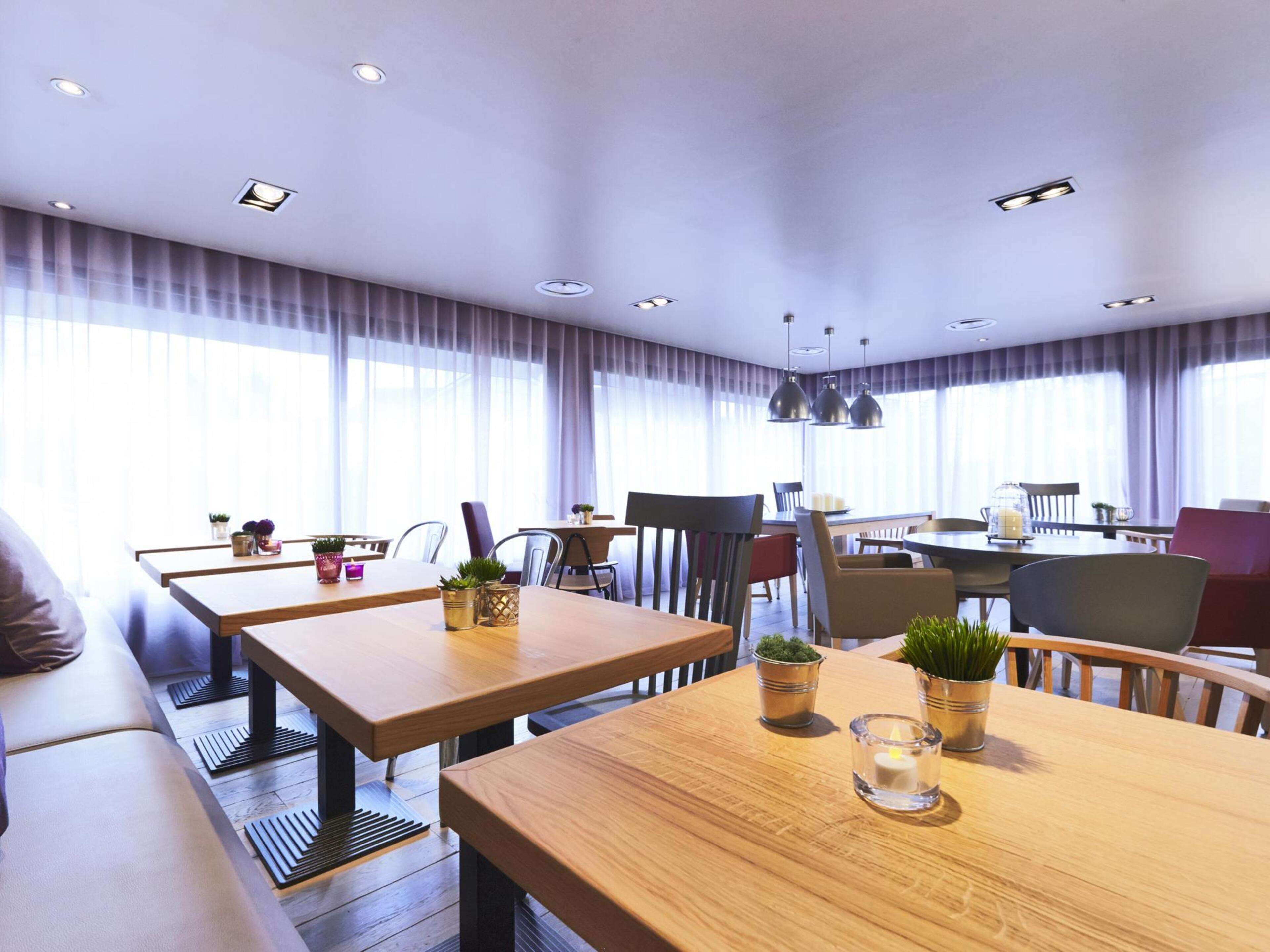 A modern dining area with wooden tables and chairs, potted plants, and pendant lighting.
