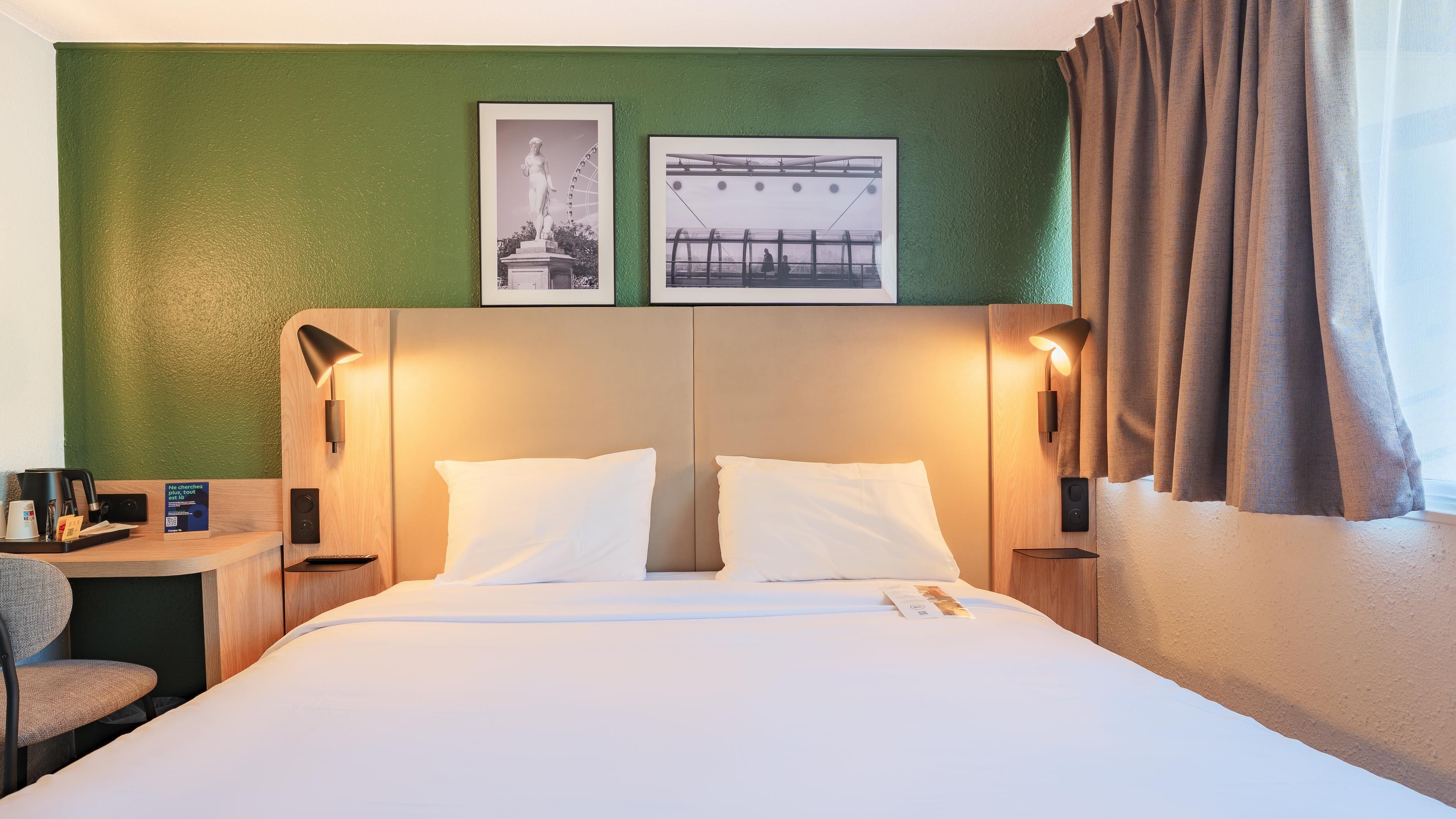 A hotel room with a bed, two wall-mounted lamps, a desk with a chair, and two framed pictures on the wall.