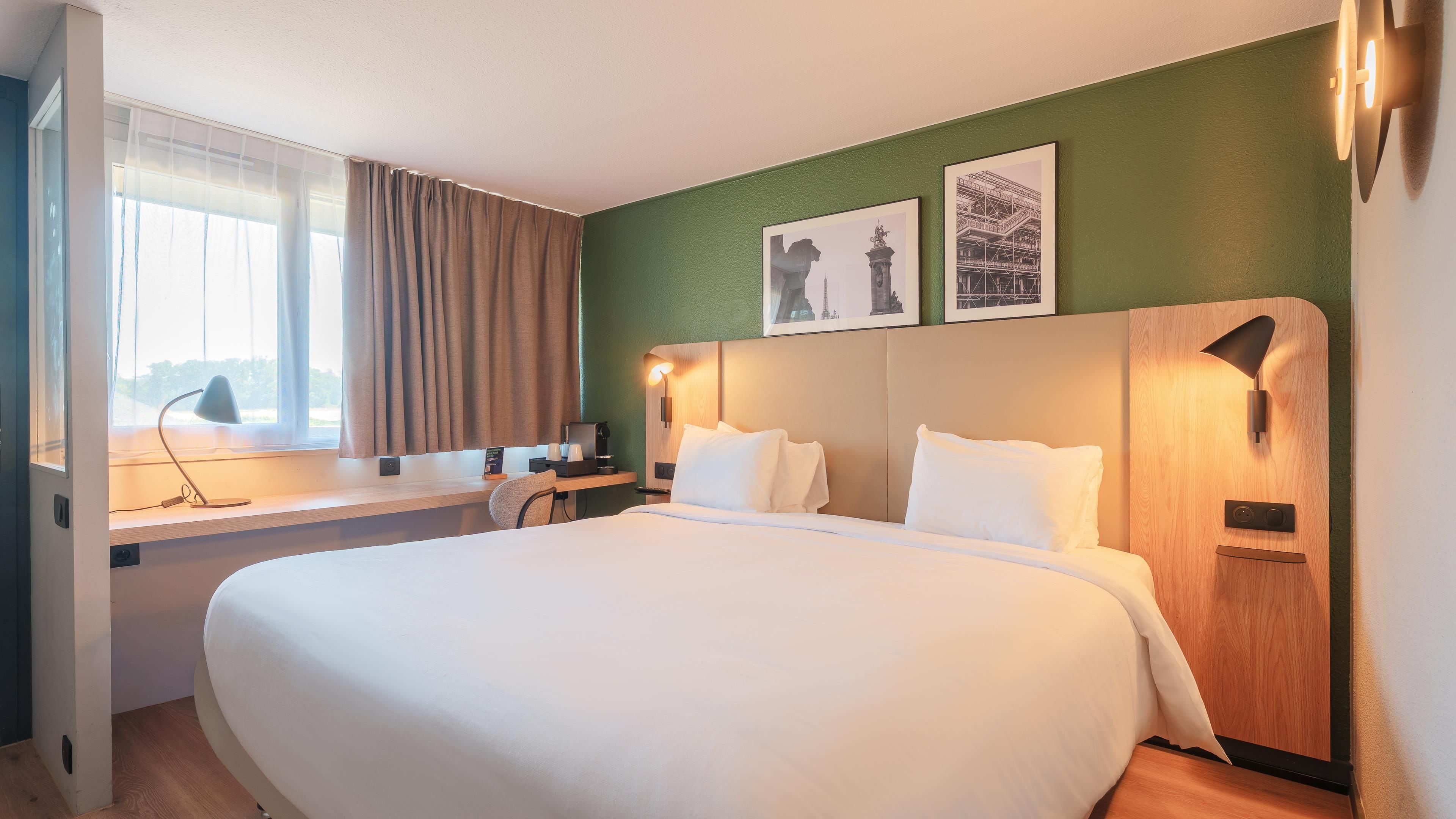 A hotel room with a large bed, a desk with a lamp, a window with curtains, and two framed pictures on the wall.