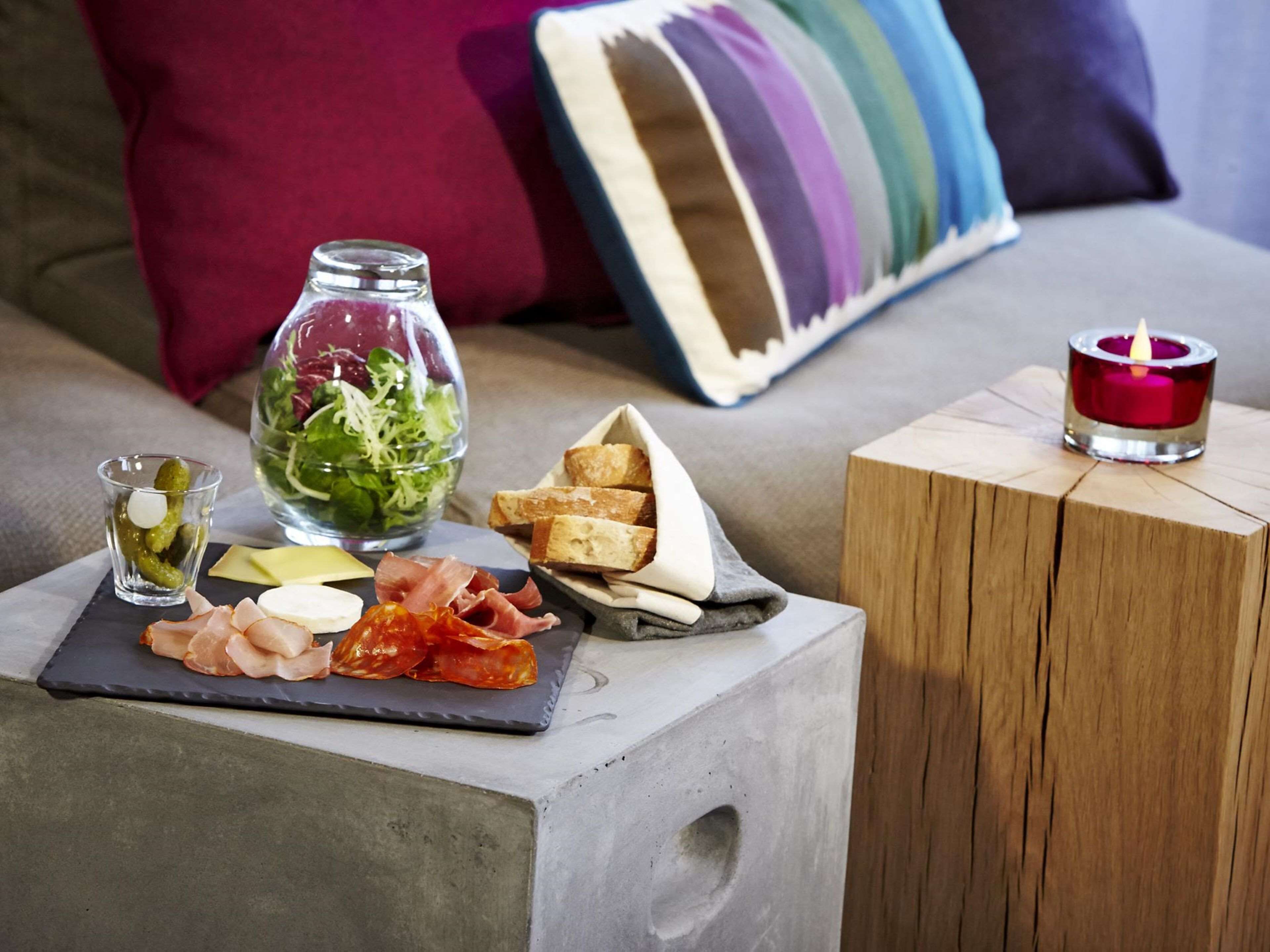 A small table with a cheese board, a glass jar of pickles, a bowl of salad, and a candle on a wooden block.