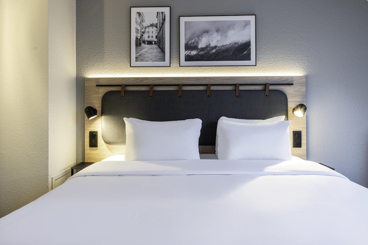 Ein modernes Hotelzimmer mit einem großen Bett, zwei Nachttischen mit Lampen, einer Kopfteilbeleuchtung und zwei gerahmten Bildern an der Wand.