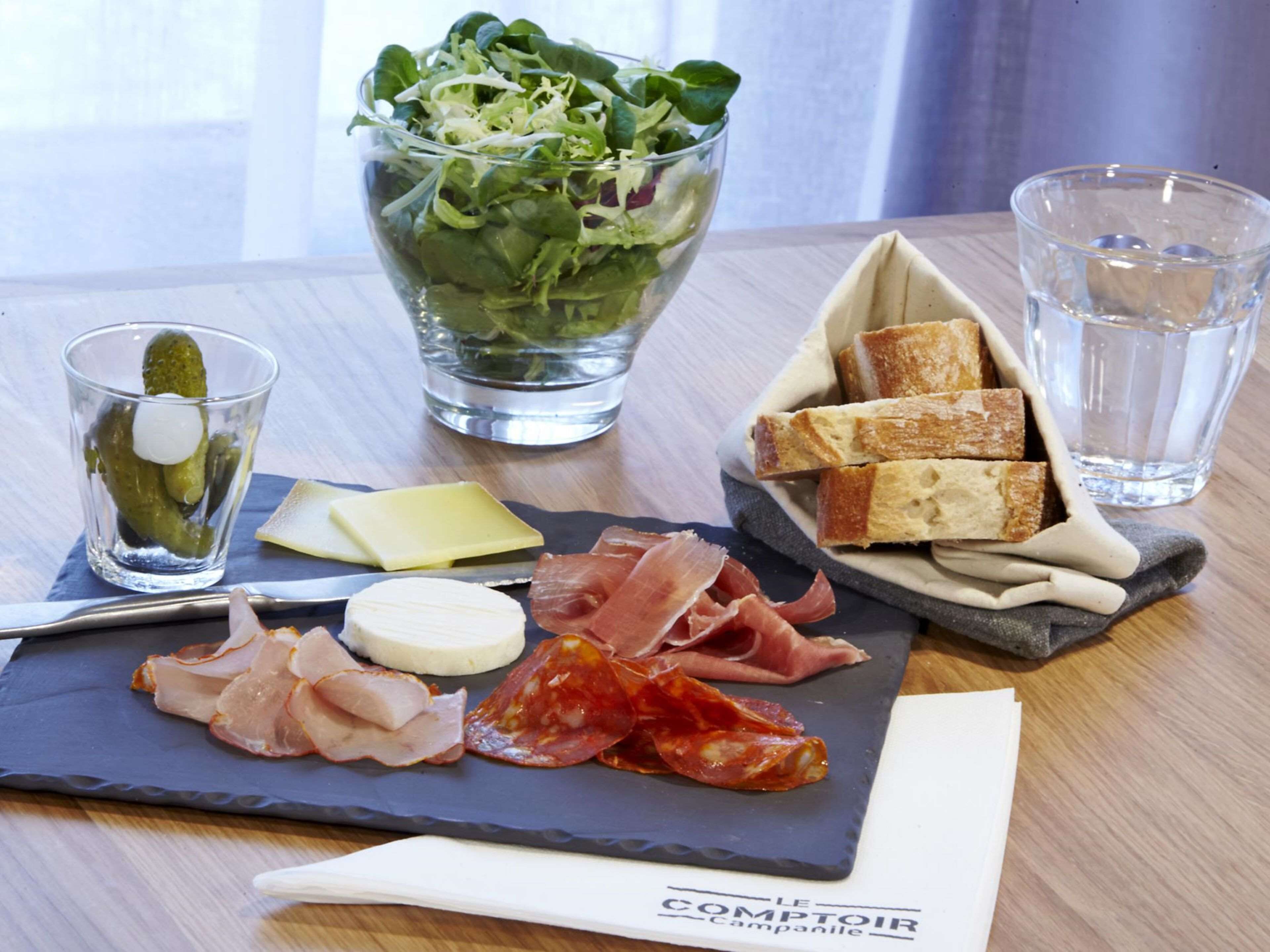 Una mesa con diversos alimentos, como ensalada, queso, embutidos y pan, junto con vasos de agua y un pepinillo.