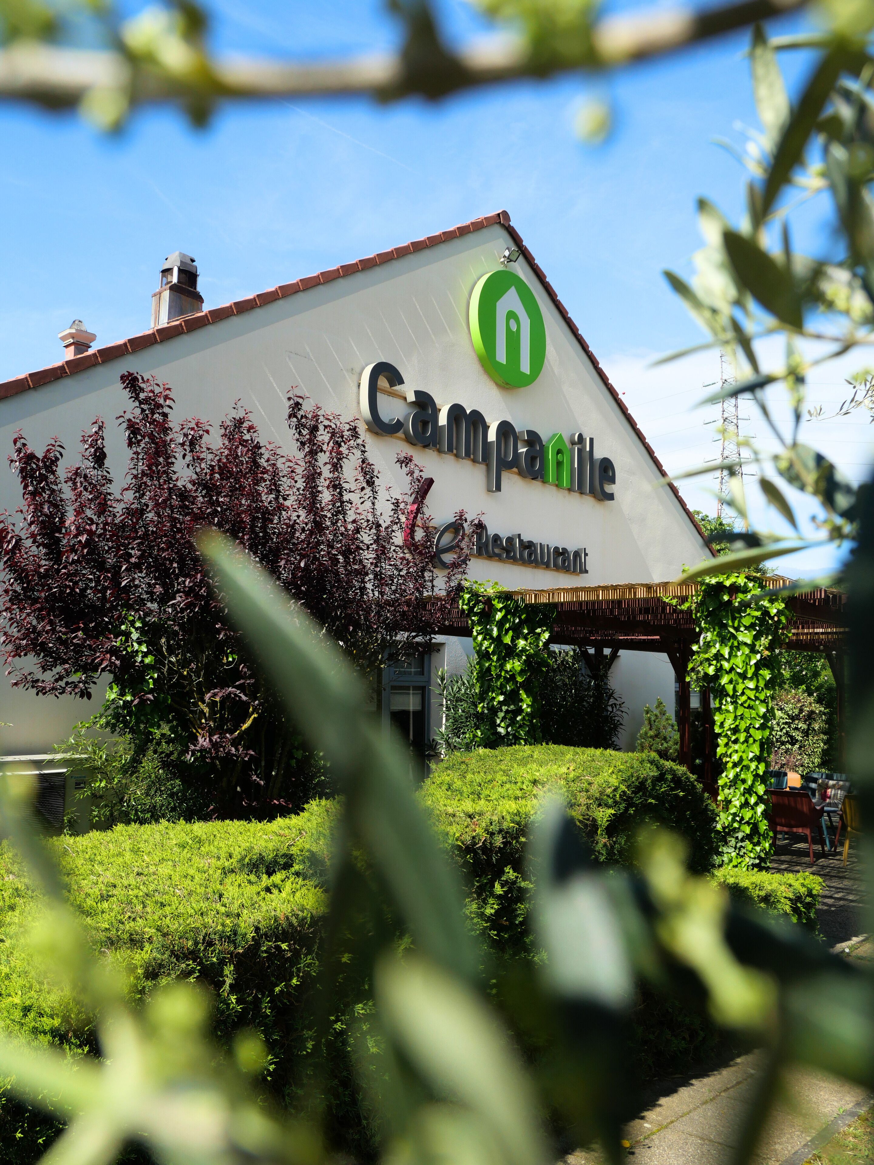 Um restaurante chamado Campanile com um logotipo verde, rodeado por vegetação e um céu claro.