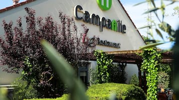 Um restaurante chamado Campanile com um logotipo verde, rodeado por vegetação e um céu claro.