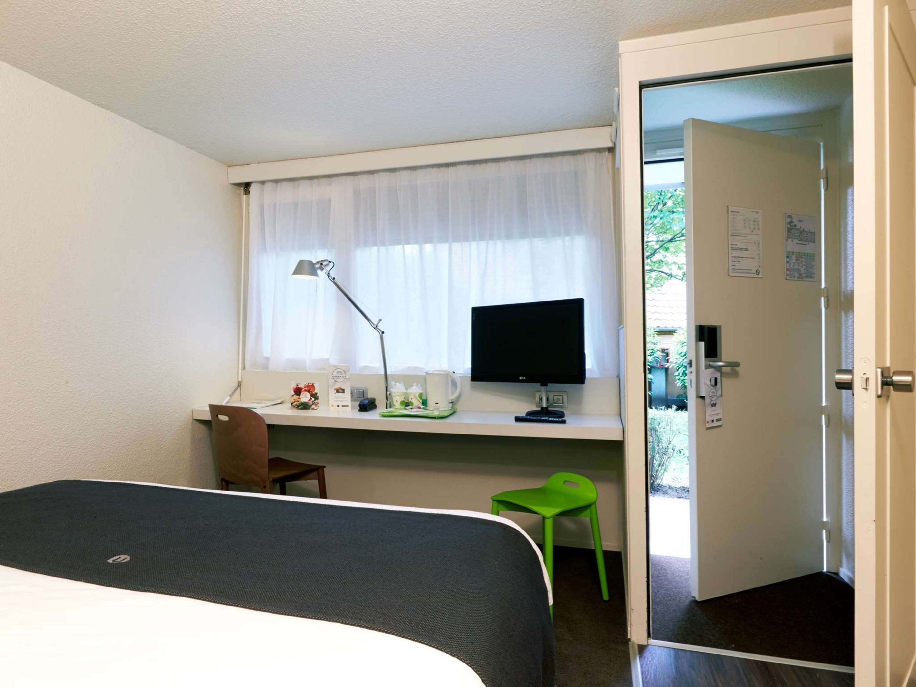 A hotel room with a bed, a desk with a computer monitor, a lamp, and a chair.