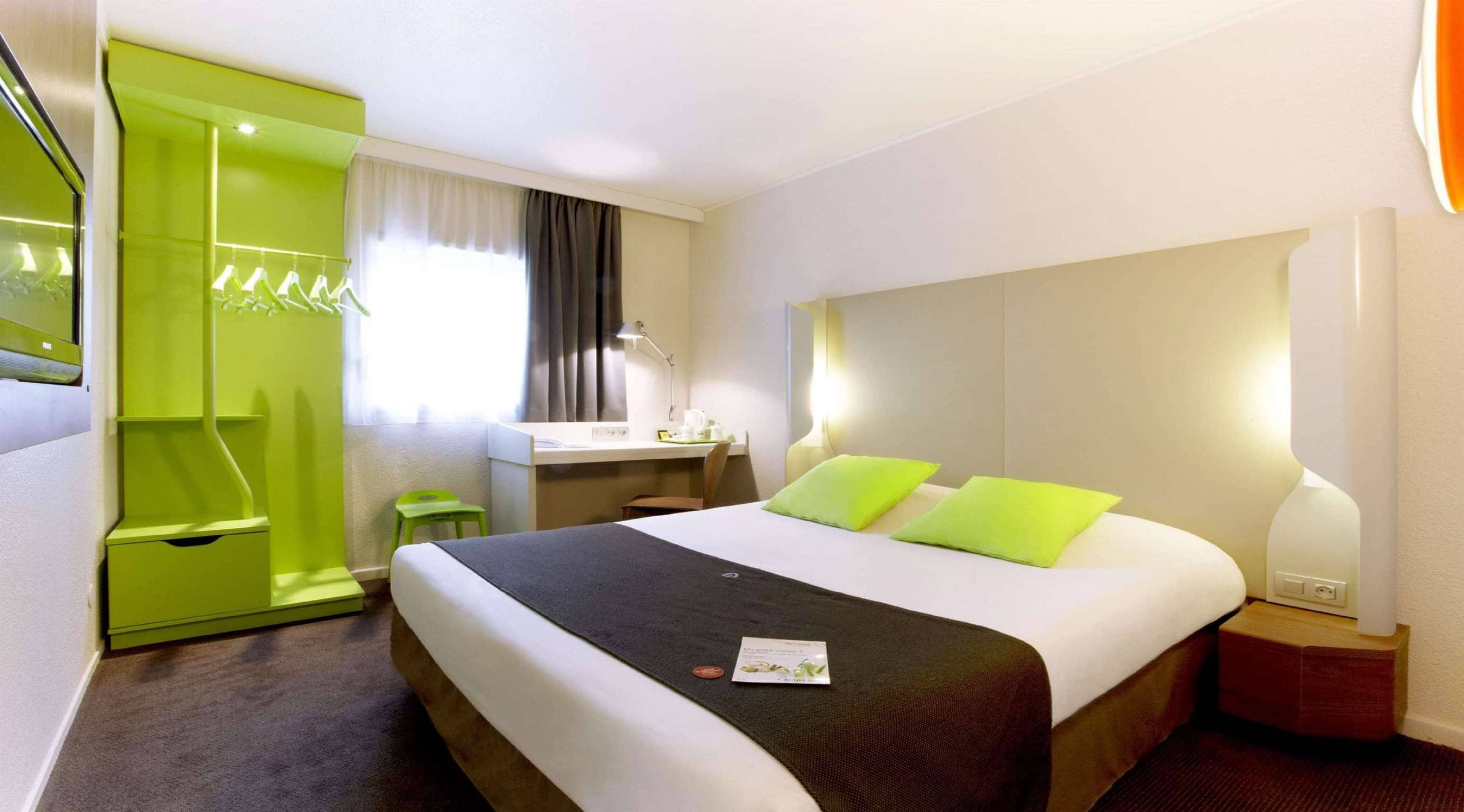 Quarto de hotel com cama, mesa de cabeceira, abajur e uma cadeira verde.