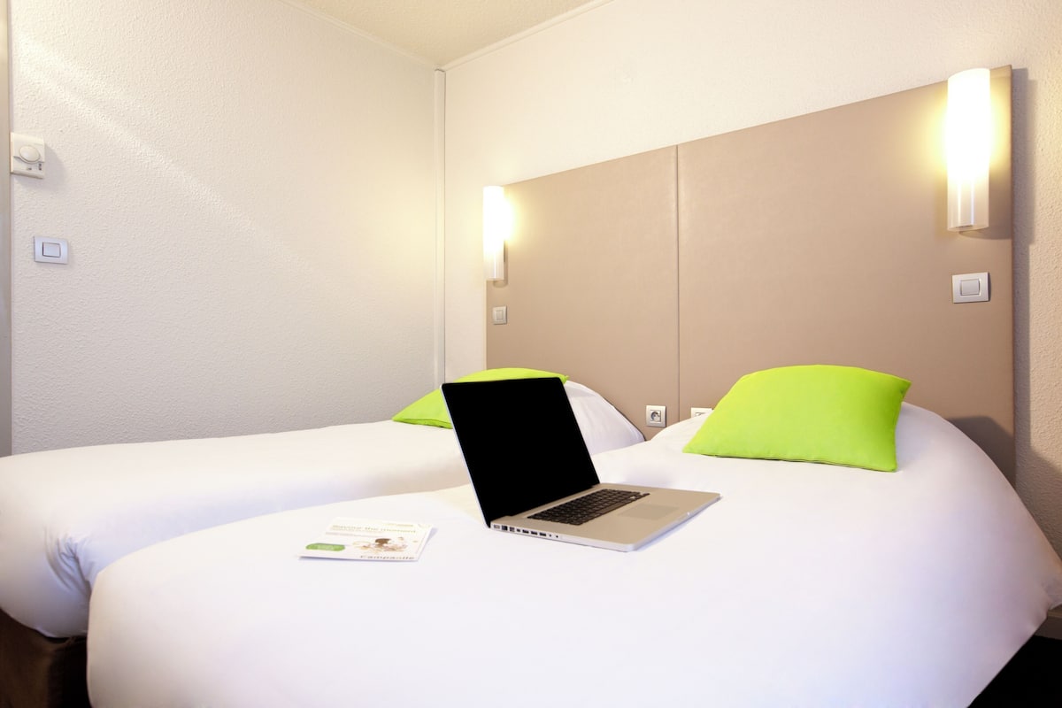 A hotel room with two beds, each with a green pillow, a laptop on one bed, and a light switch on the wall.