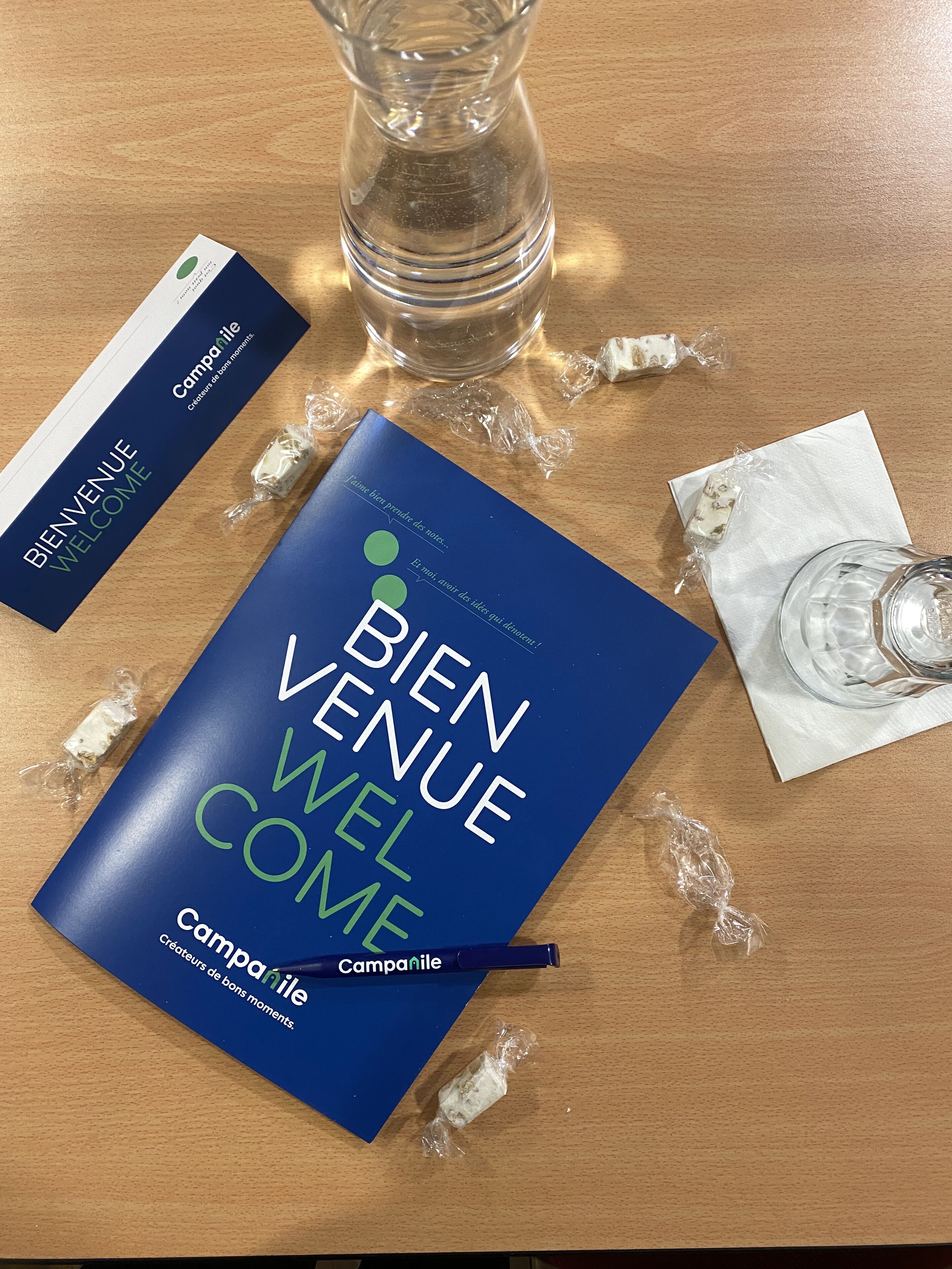 A welcome booklet, a pen, a glass, and some wrapped items on a wooden table.