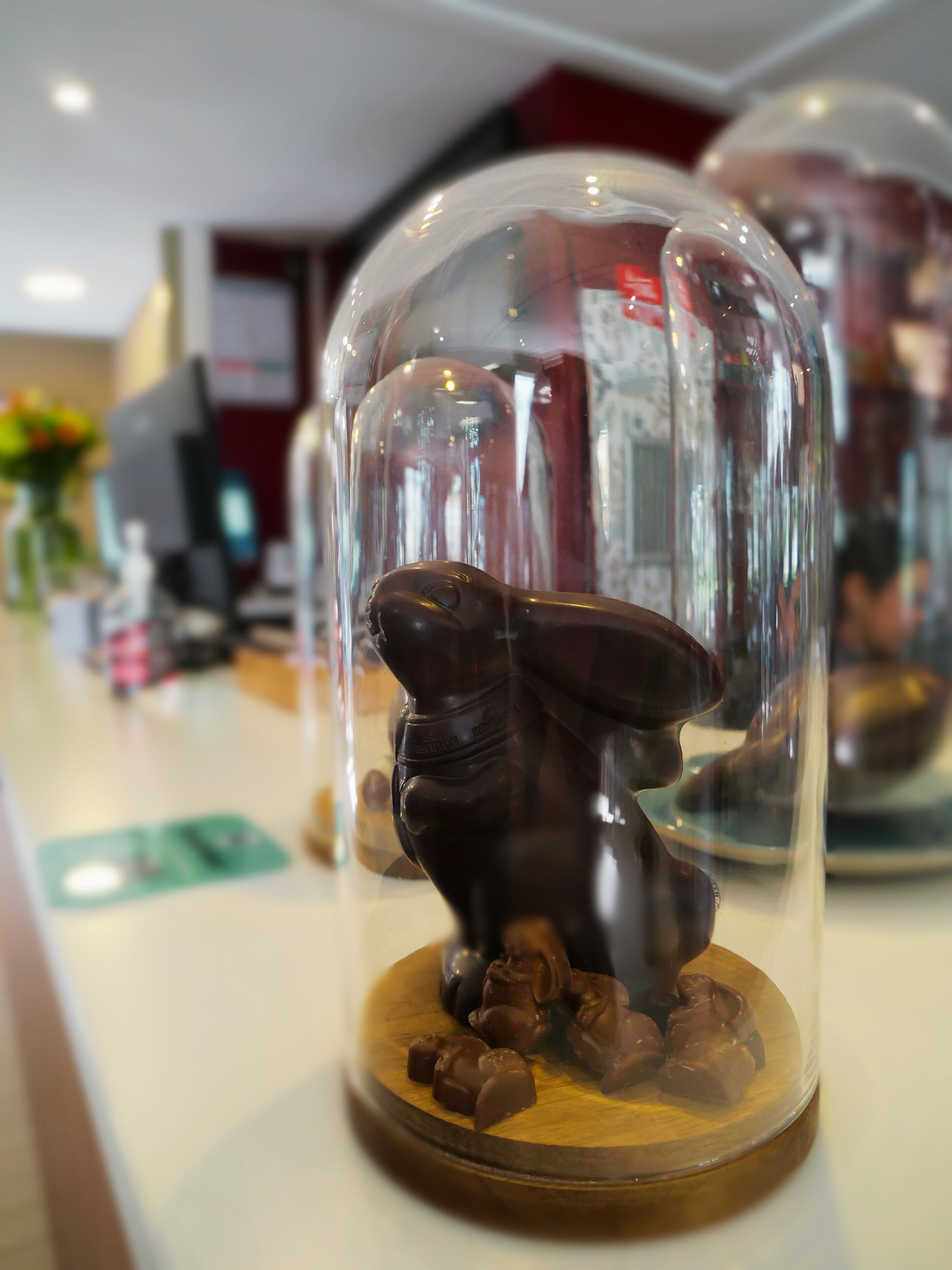 Une sculpture en chocolat recouverte de chocolat, placée sous une cloche de verre, avec un arrière-plan flou représentant l’intérieur d’un restaurant.