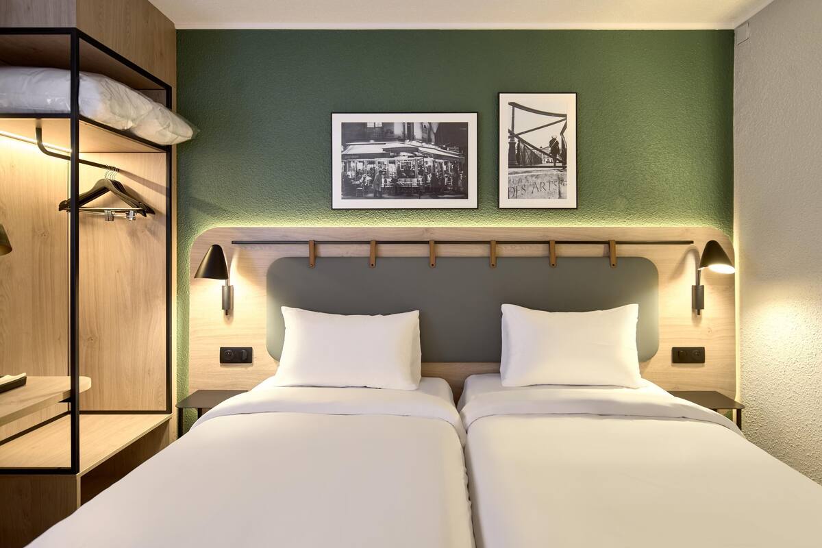 A hotel room with two beds, a wooden wardrobe, and two framed pictures on the wall.