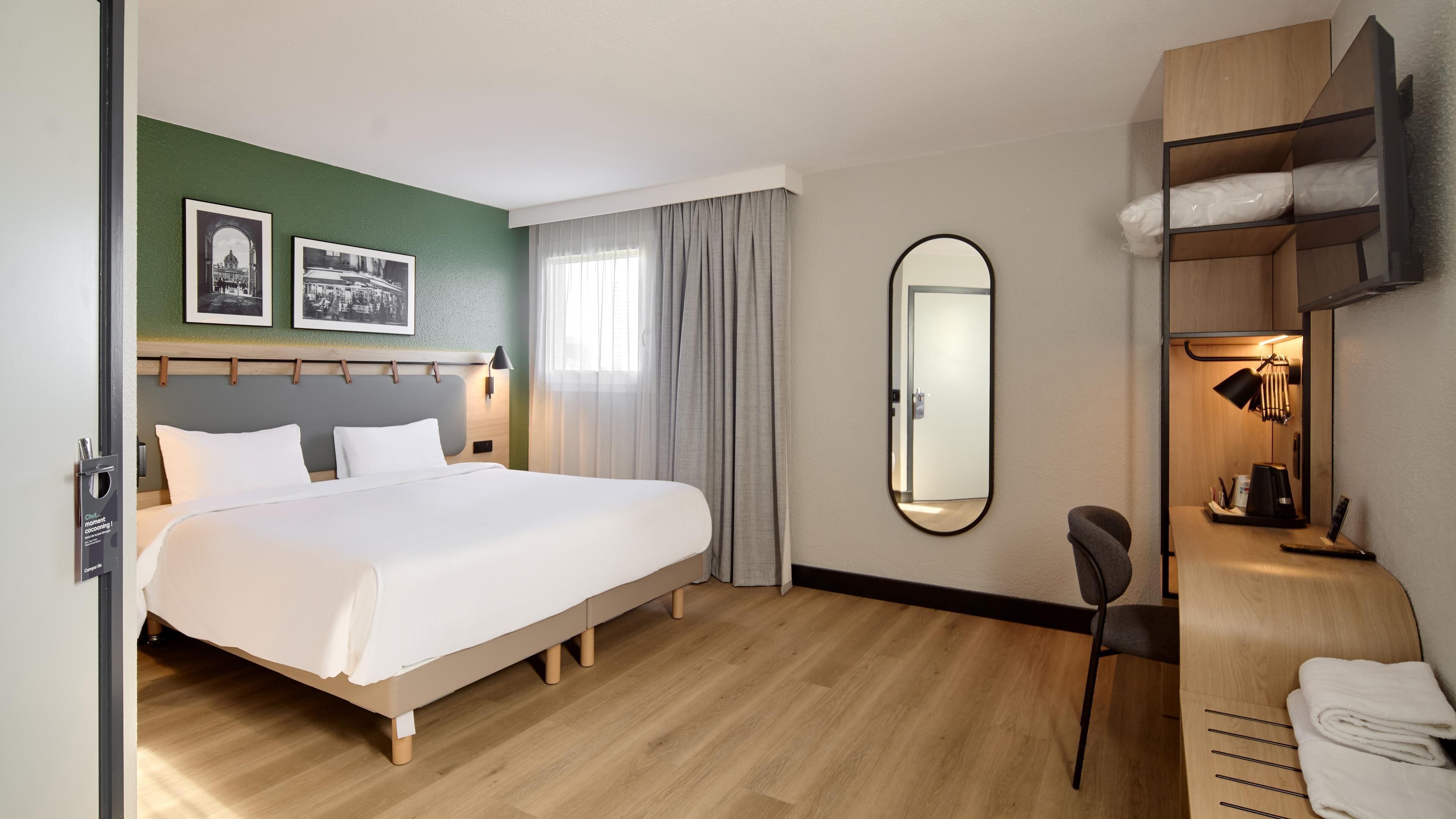 A hotel room with a bed, a desk with a chair, a mirror, and a wardrobe.