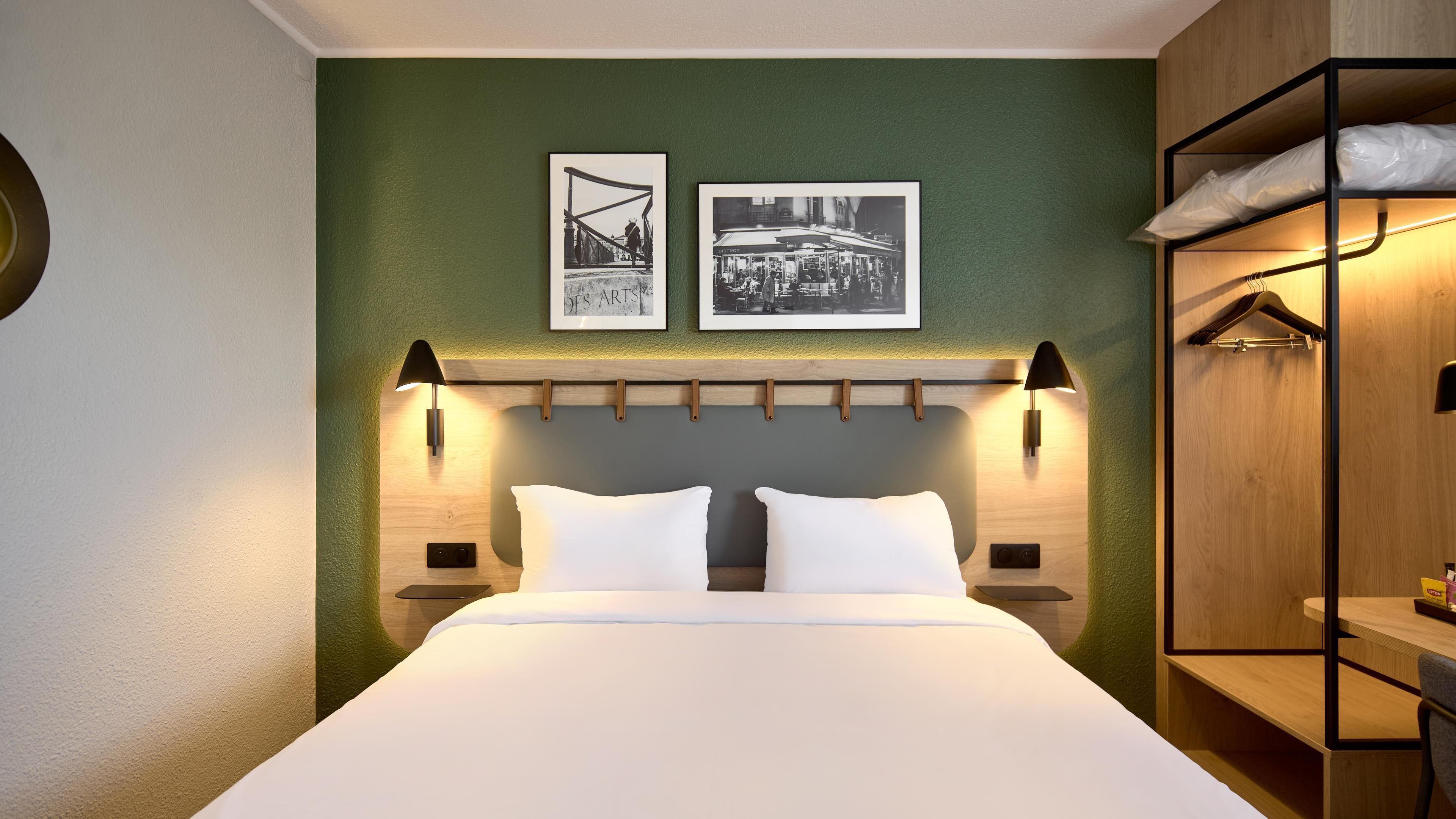 A modern hotel room with a large bed, two wall-mounted lamps, a wooden headboard, and two framed pictures on the wall.