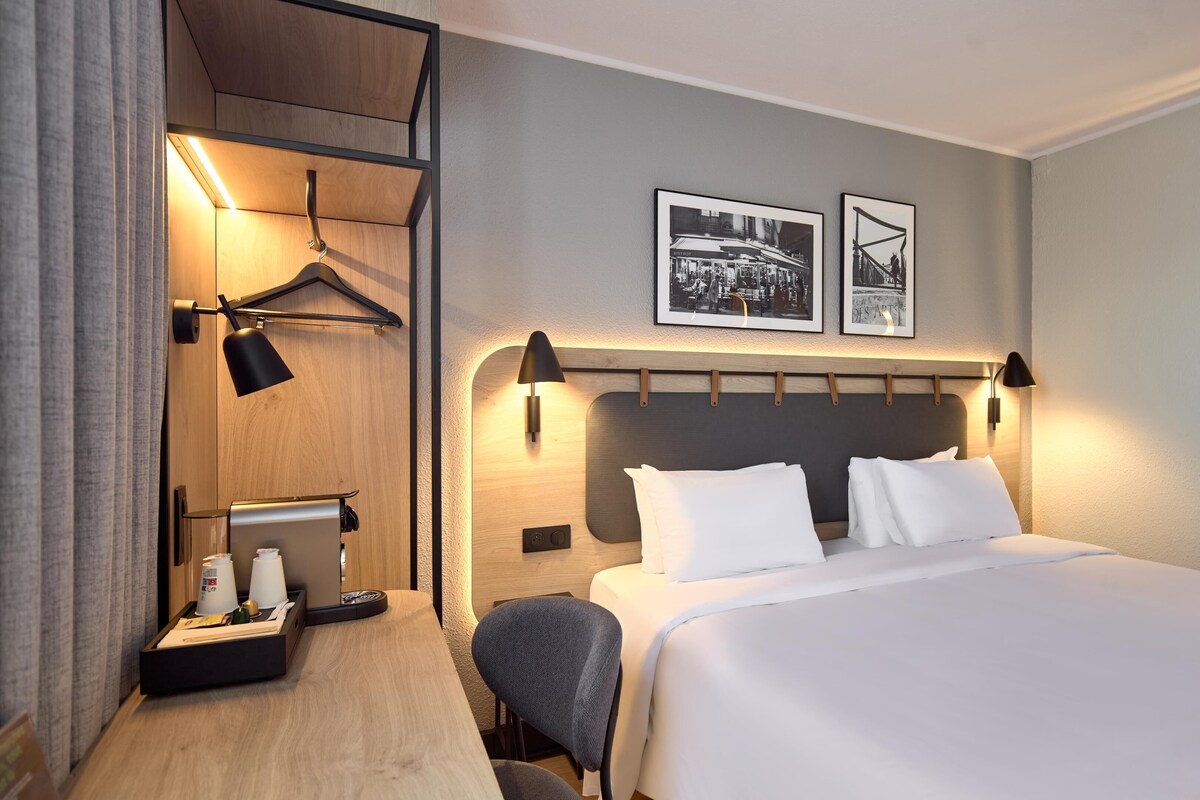 A hotel room with a bed, a desk with a computer, a chair, a coat rack, and two framed pictures on the wall.