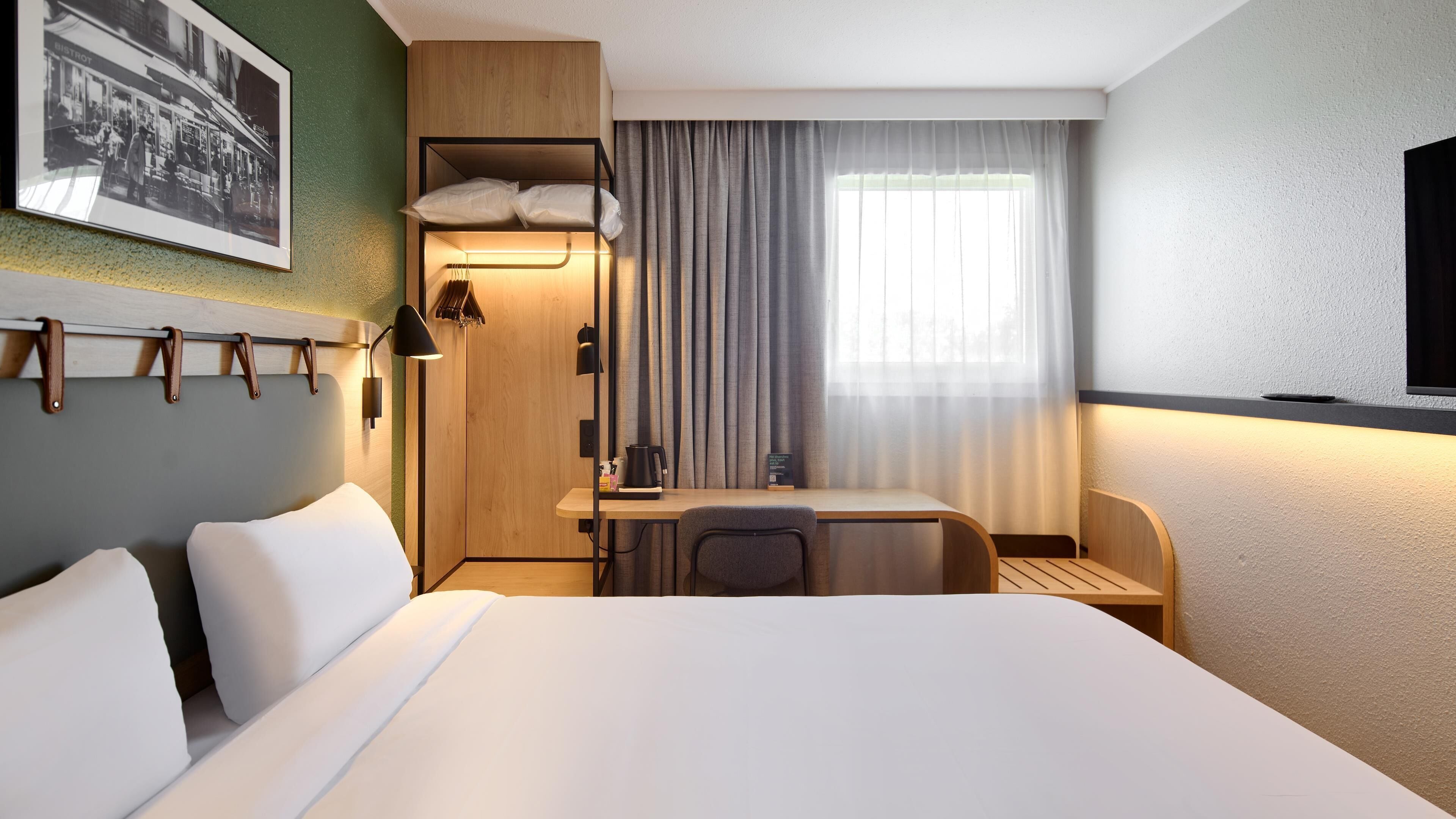 A hotel room with a large bed, a desk with a chair, a small table with a kettle, and a television.