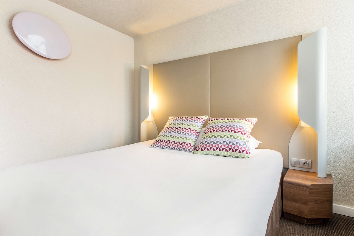 Una cama bien hecha con almohadillas estampadas, una lámpara de noche y un espejo montado en la pared.