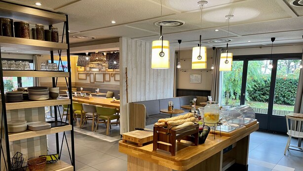 A modern cafe with a central wooden counter, shelves with glass jars, and a view of greenery outside.