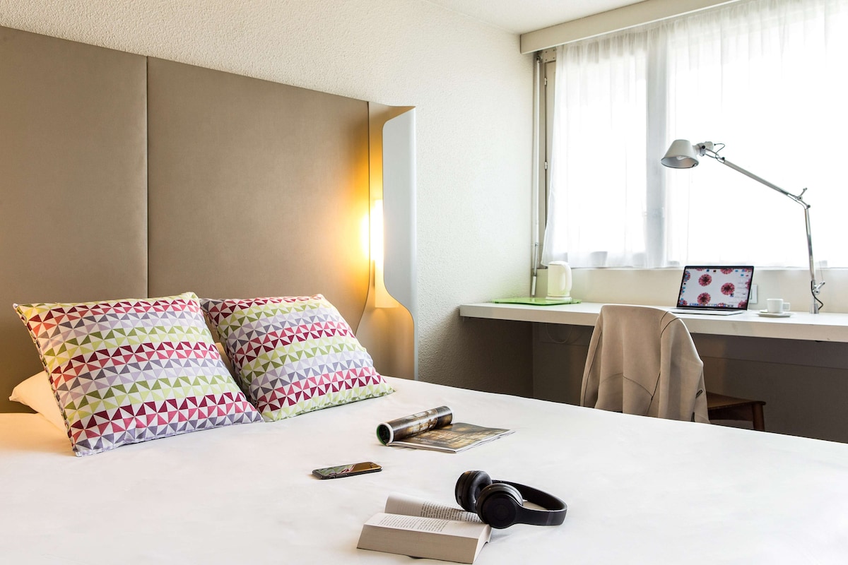 Een netjes opgemaakt bed met patroon kussens, een nachtkastje met een lamp, een laptop en een stoel.