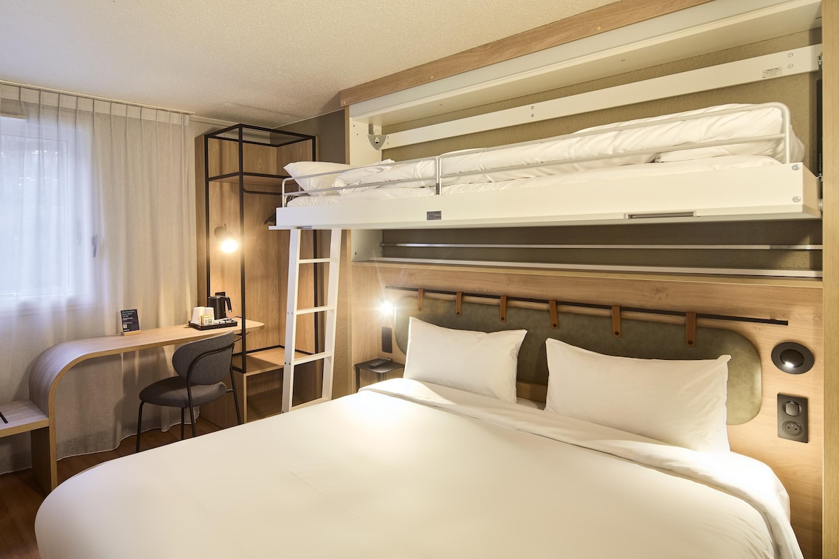 Habitación con literas, escritorio y silla, una mesita con cafetera y ventana con cortinas.