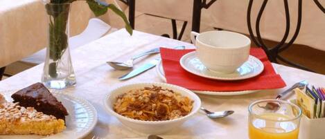 Una escena de desayuno con una mesa preparada para dos personas, que incluye platos, tazones, cubiertos, un florero con una rosa y una cesta de frutas al fondo.
