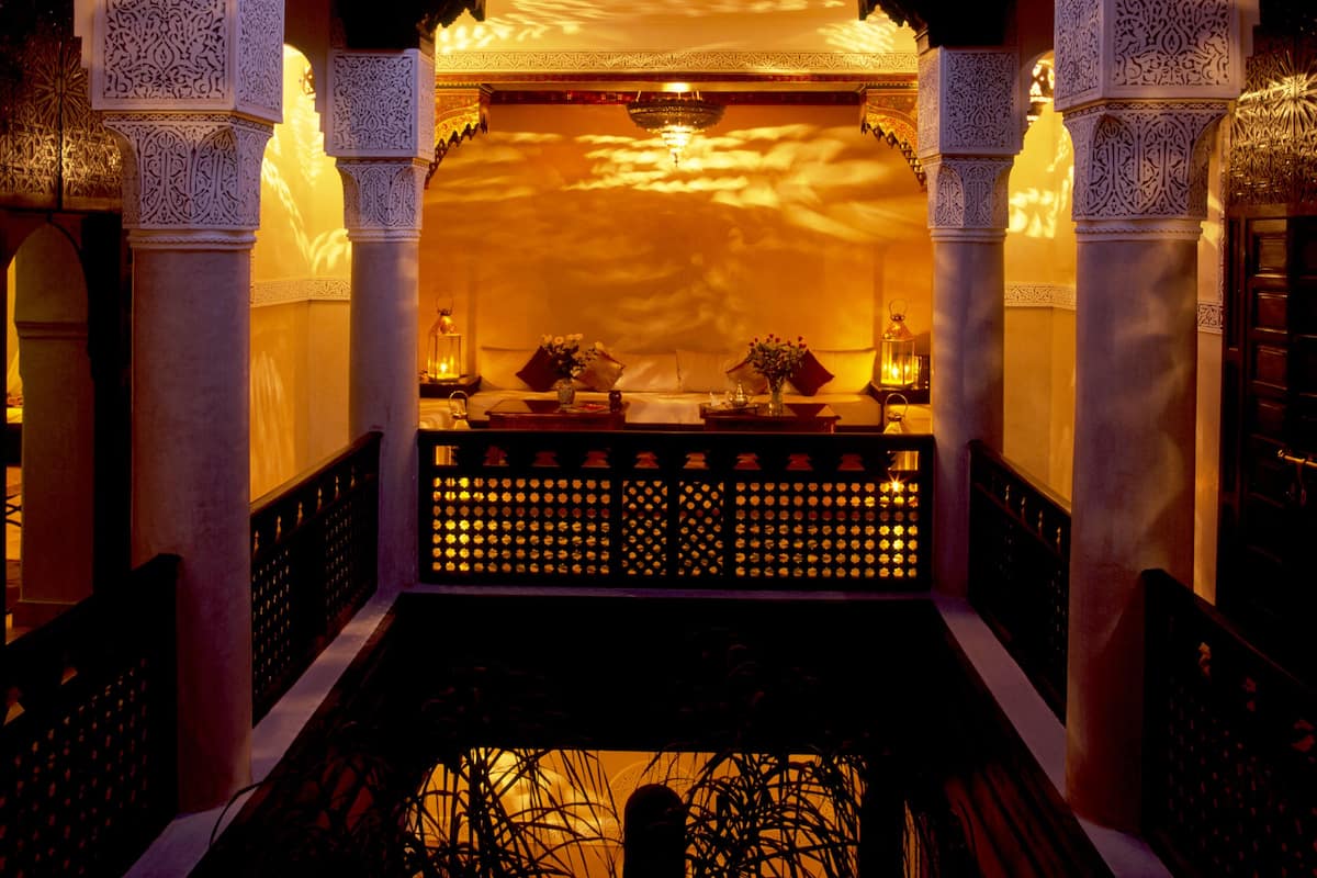 An ornate corridor with intricate architectural details, a central seating area, and a view of the sky.